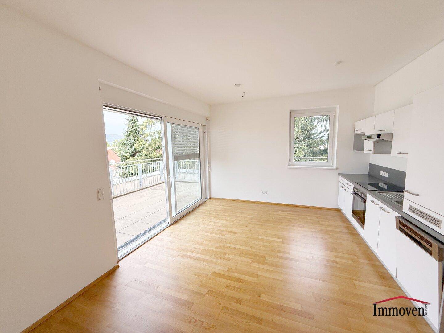 Heller Wohnbereich mit Holzboden, weißer Einbauküche und Zugang zu einer großen Terrasse.
