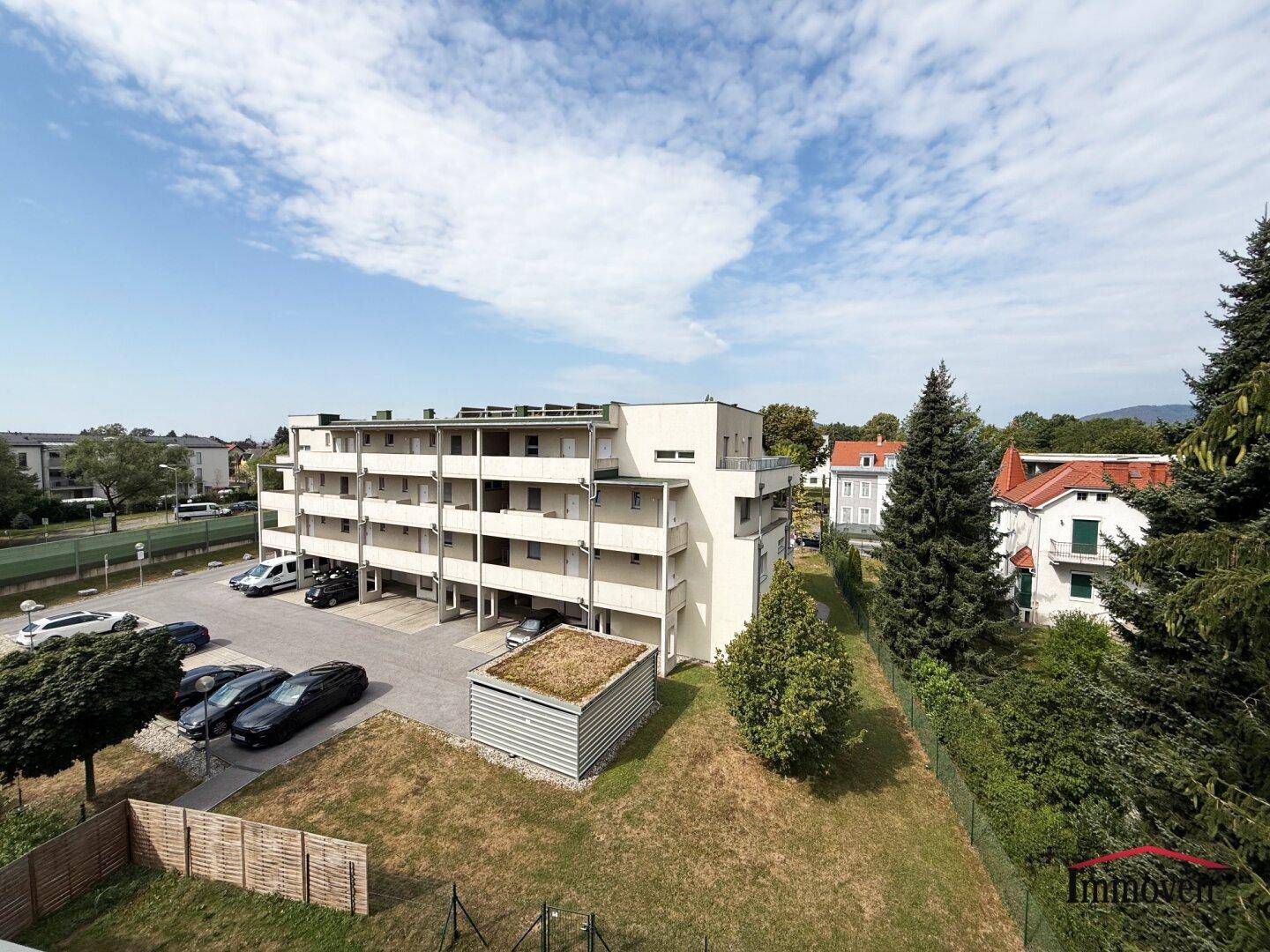 Gesamtansicht des Apartmentgebäudes mit Parkplätzen und grüner Umgebung unter blauem Himmel.