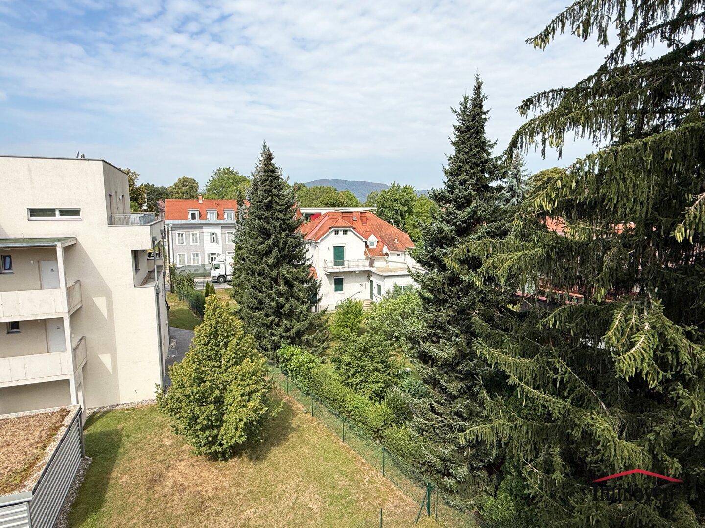 Panoramablick über grüne Bäume, umliegende Häuser und ferne Berge unter blauem Himmel.