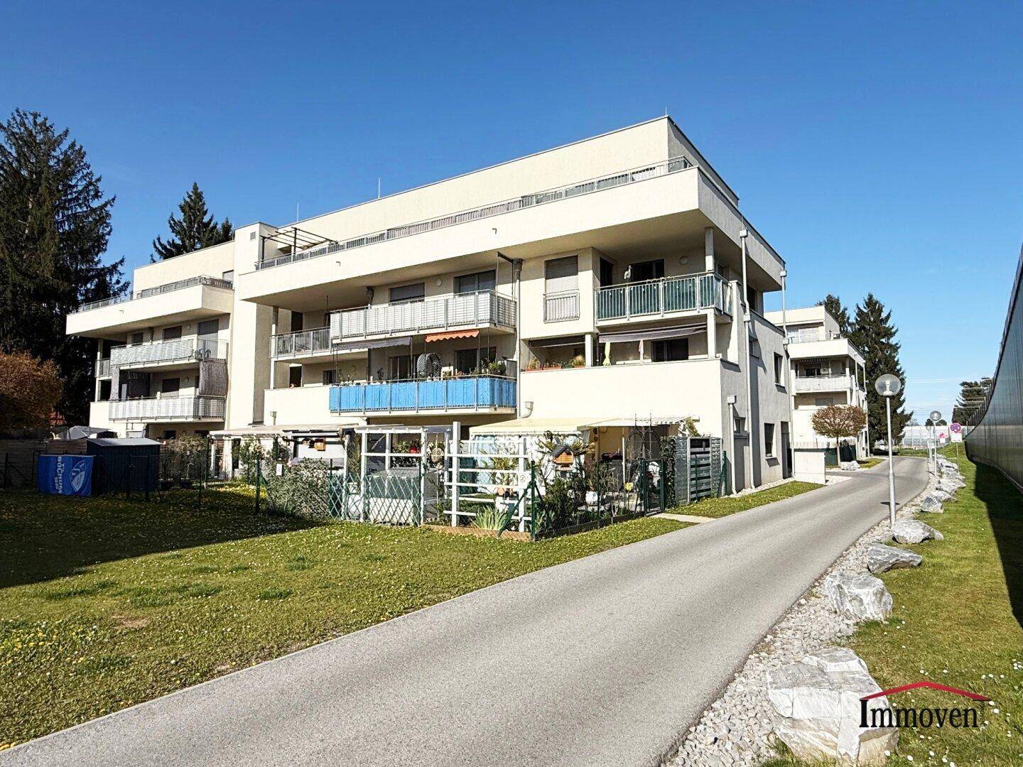 Modernes Apartmentgebäude mit mehreren Balkonen, umgeben von Grünflächen und einer Zufahrtsstraße.