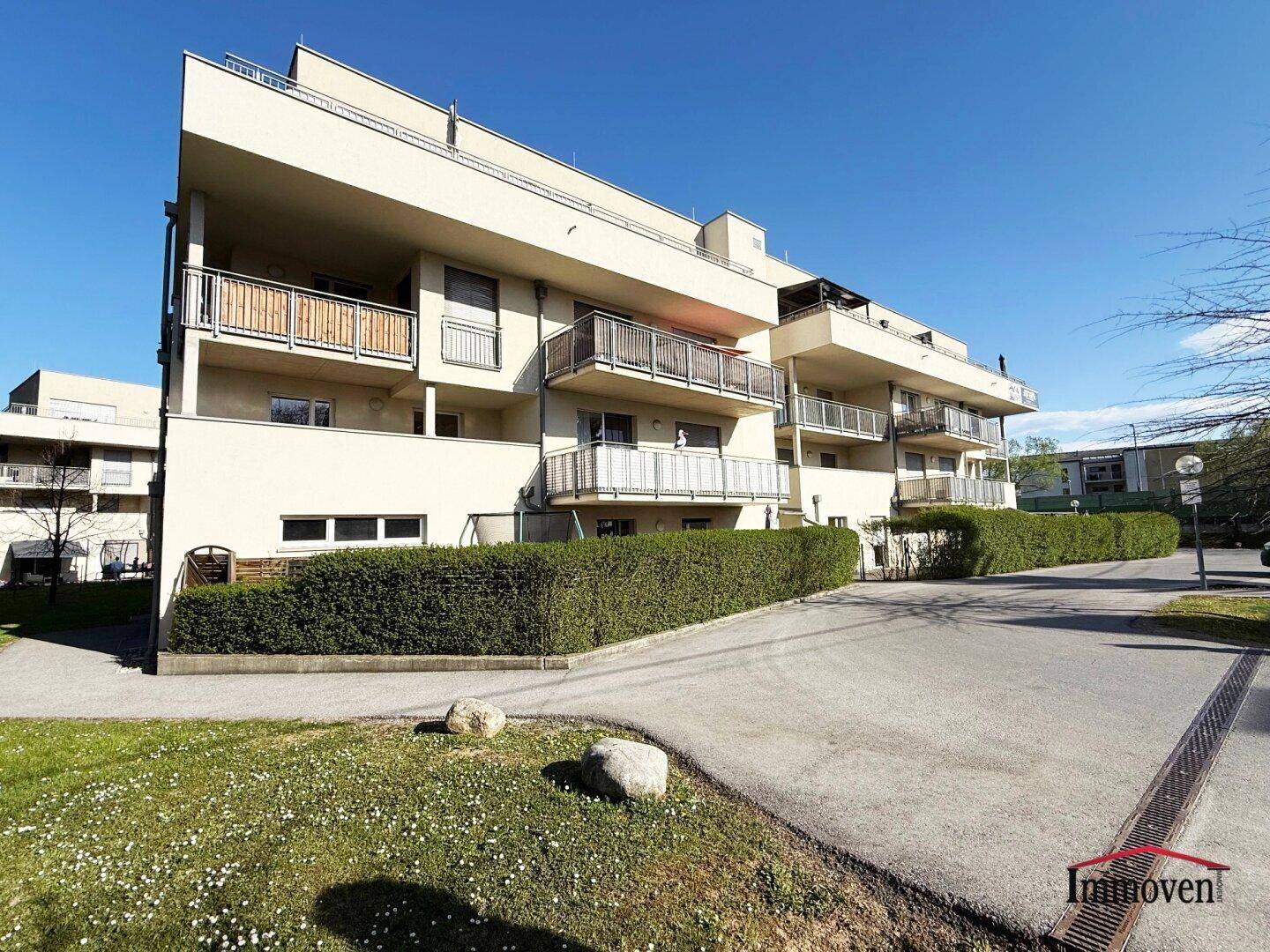 Ansicht des Apartmentgebäudes mit Balkonen, gepflegter Hecke und Zufahrt an einem sonnigen Tag.