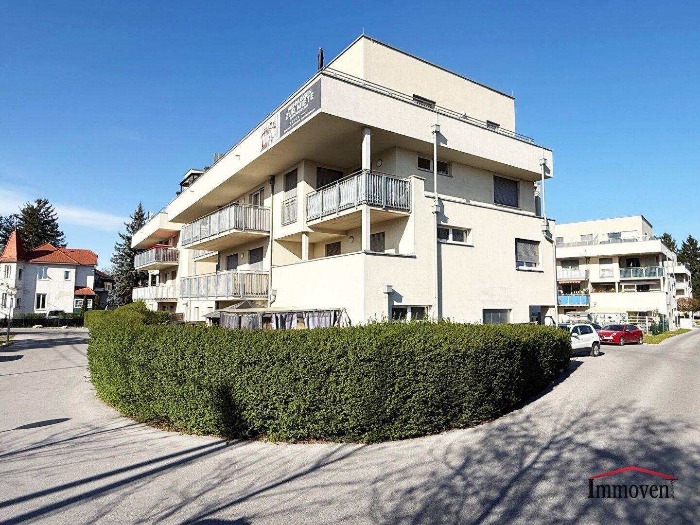 Eckansicht des modernen Apartmentgebäudes mit Balkonen und einer dichten Hecke im Vordergrund.