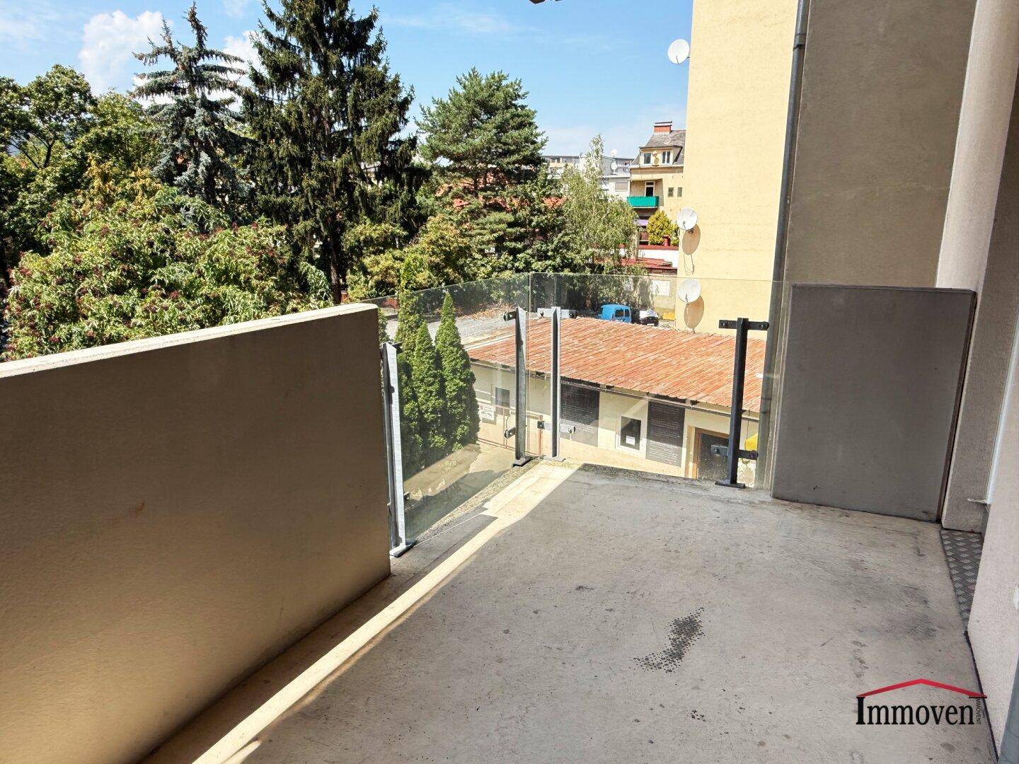 Geräumiger Balkon mit Glasgeländer und Blick ins Grüne sowie auf umliegende Gebäude.
