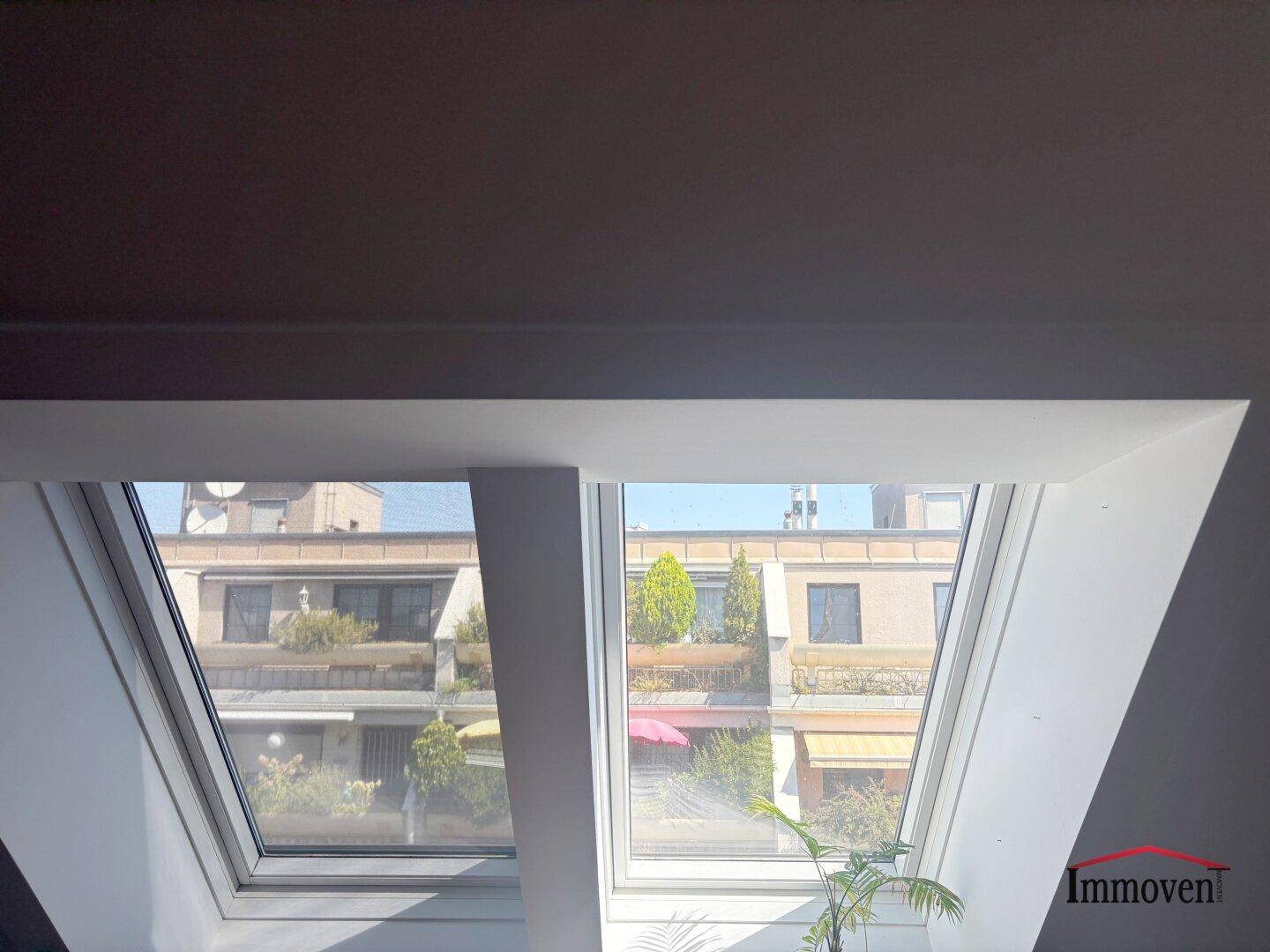 Blick durch zwei Dachfenster auf umliegende Gebäude und grüne Bäume.
