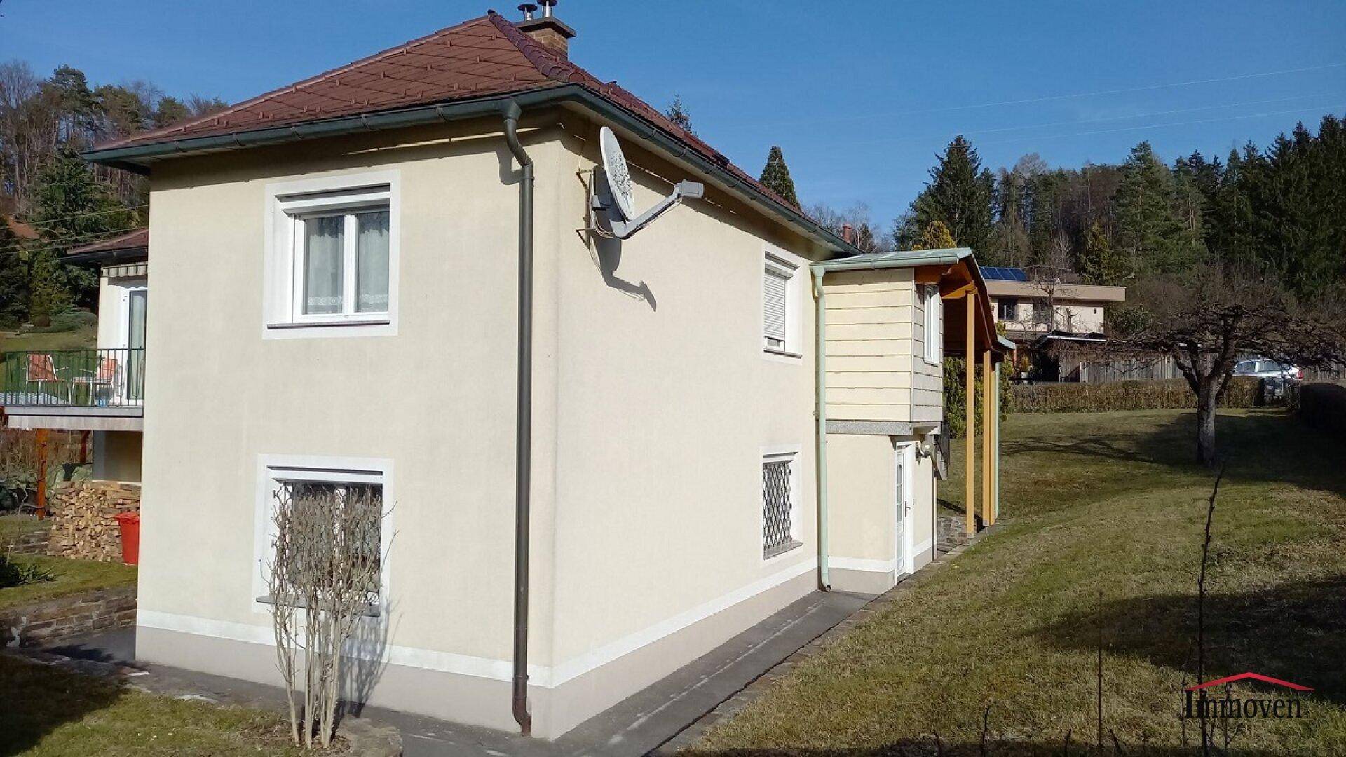 Helles Einfamilienhaus mit beiger Fassade, braunem Ziegeldach und gepflegtem Gartenbereich unter blauem Himmel.