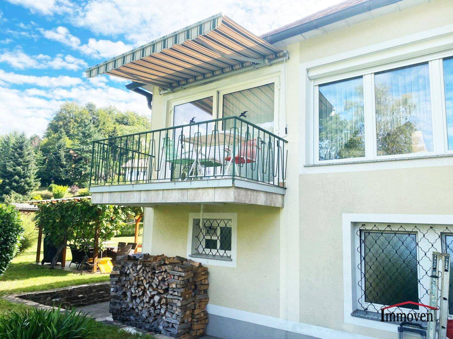Balkon mit Markise und Metallgeländer, darunter ein Holzstapel im Gartenbereich.