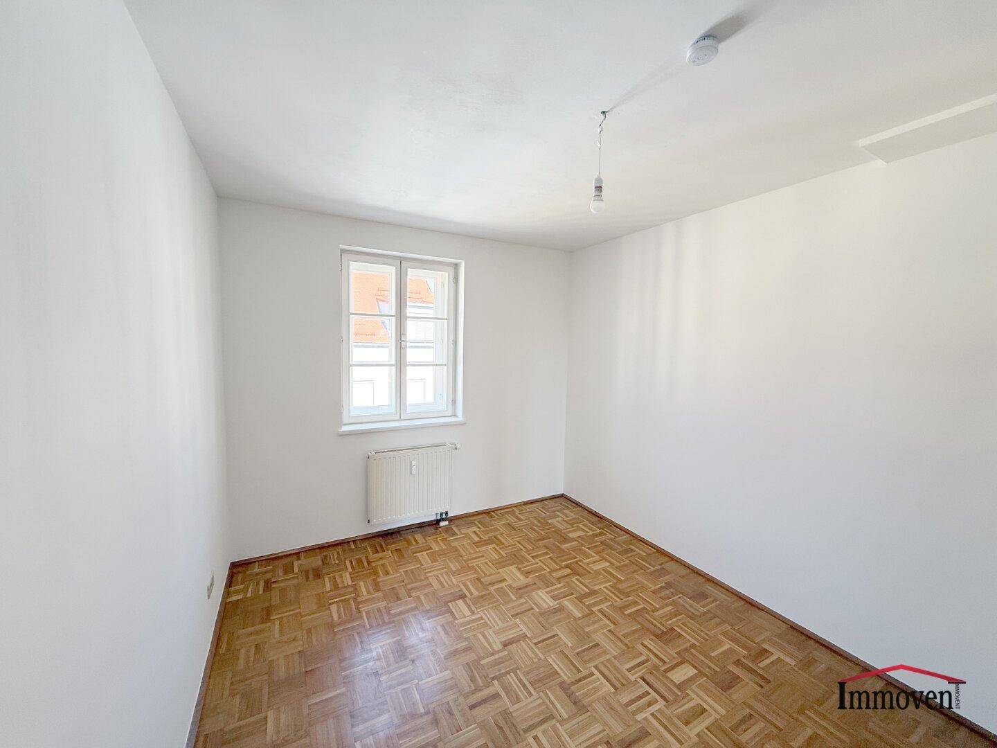 Schlichtes Schlafzimmer mit Parkettboden, weiß gestrichenen Wänden und einem Fenster.