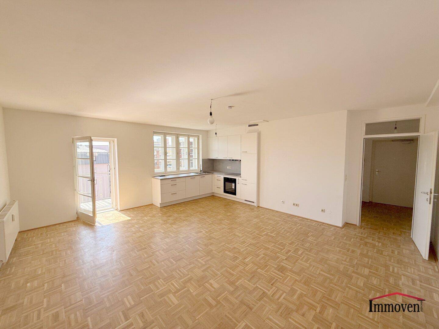 Heller Wohnbereich mit Parkettboden, integrierter Küchenzeile und Zugang zum Balkon.