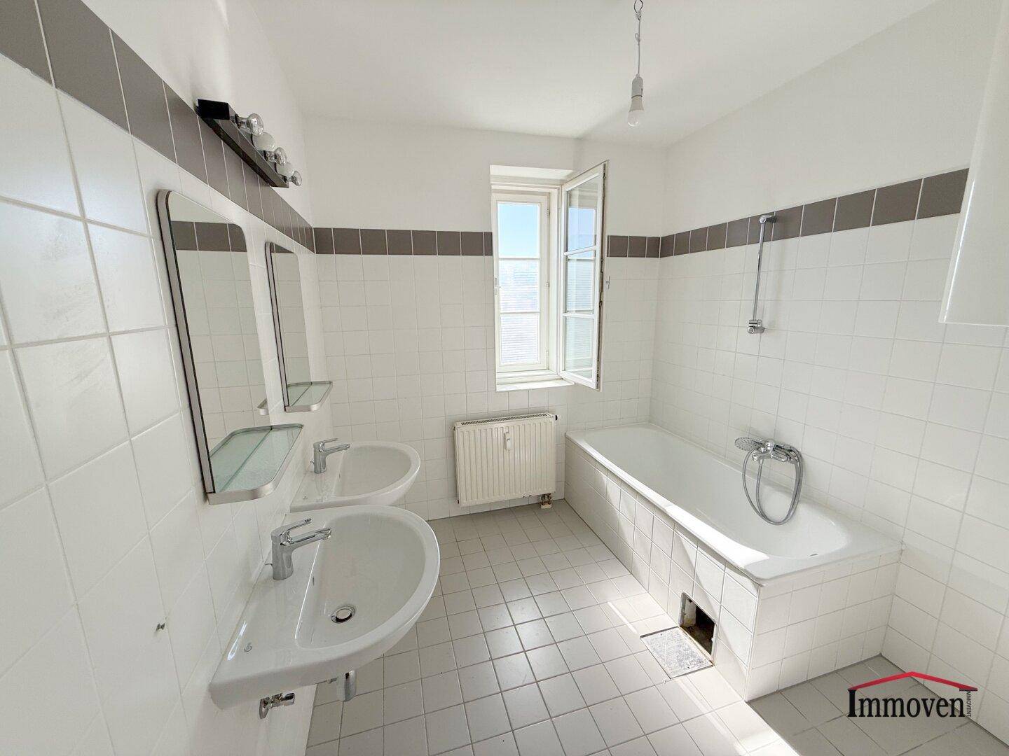 Badezimmer mit Badewanne, Doppelwaschbecken und Fenster, gefliest in Weiß und Grau.