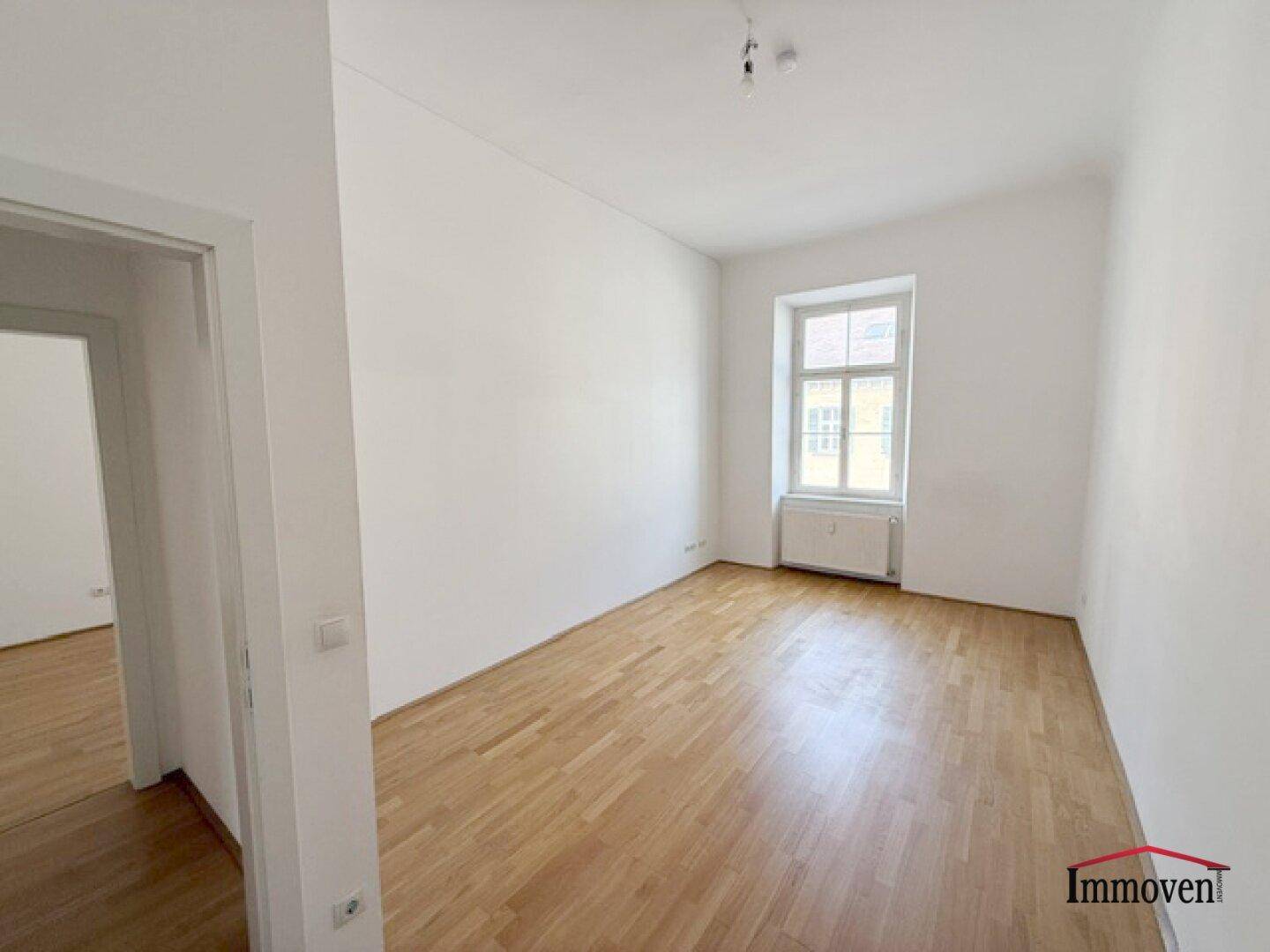 Geräumiges Zimmer mit hellem Parkettboden und einem großen Fenster, das viel Tageslicht hereinlässt.