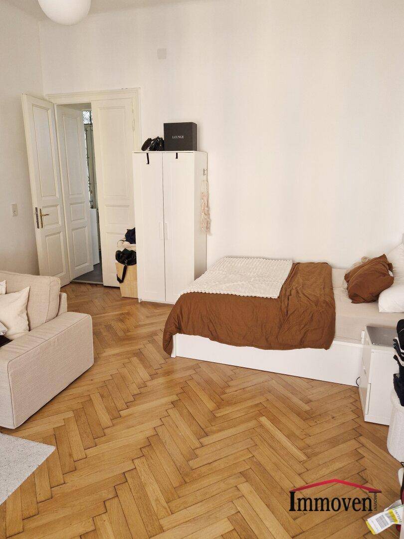 Schlafzimmer mit beigem Bett, weißem Kleiderschrank und klassischem Fischgrätparkett.