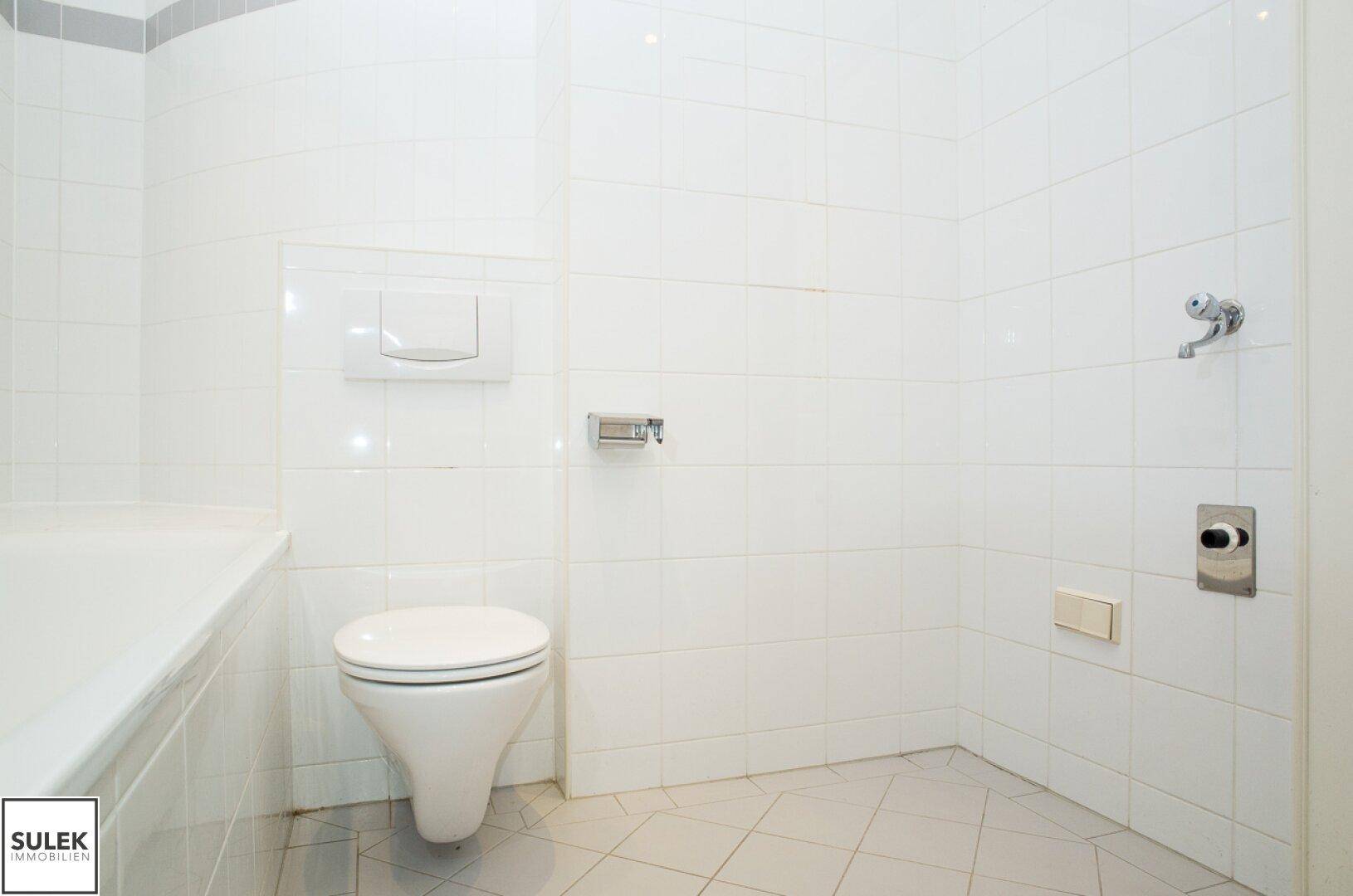Heller Badezimmerbereich mit weißen Fliesen, wandhängendem WC und Platz für eine Dusche in der Ecke.