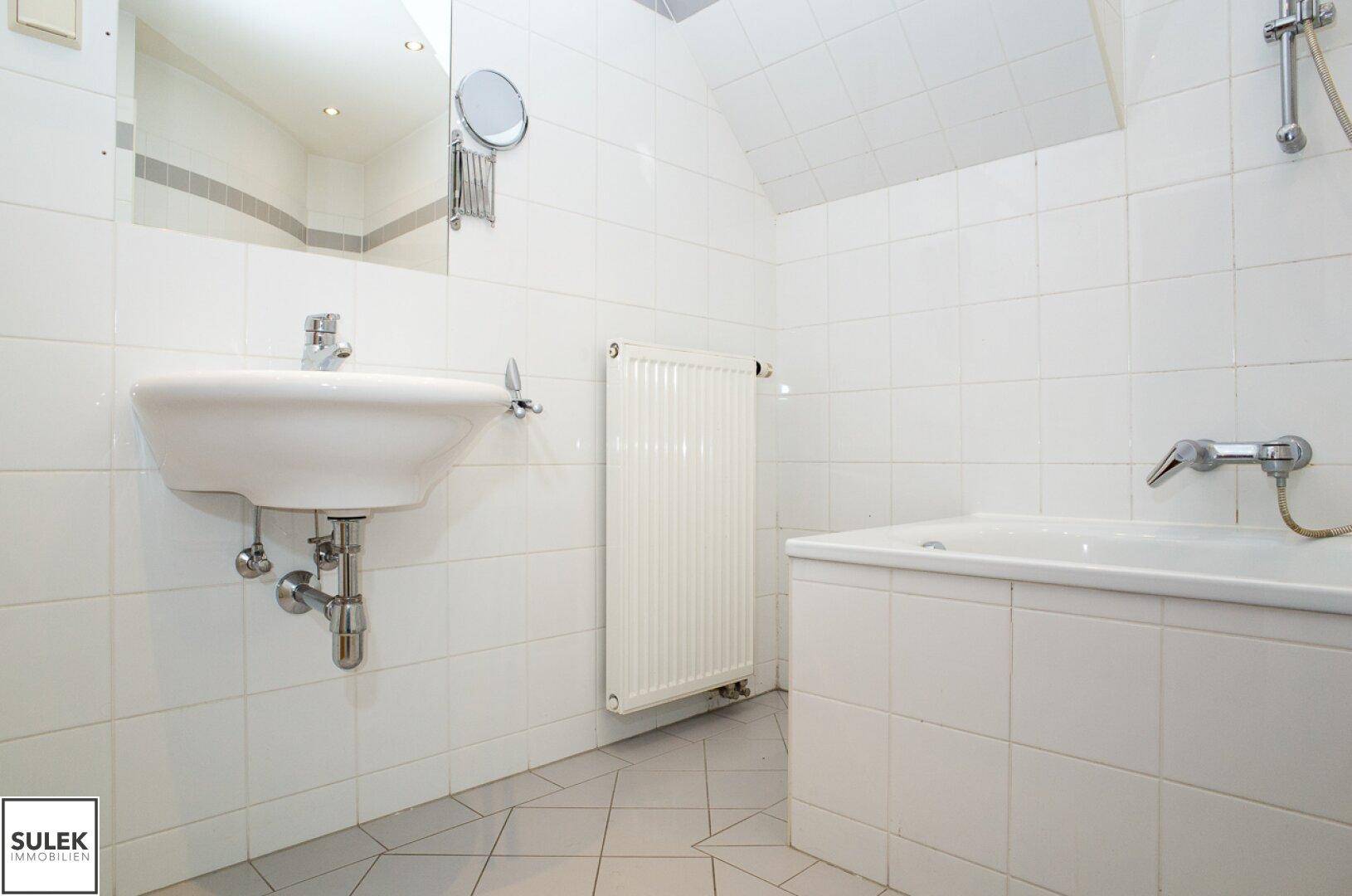 Badezimmer mit Waschbecken, Spiegel und Heizkörper, komplett gefliest in Weiß und Grau.