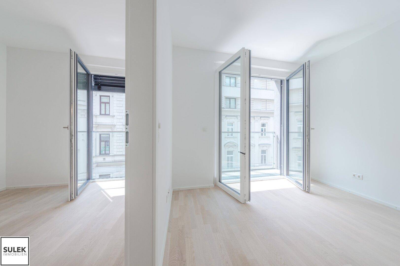 Lichtdurchflutetes Zimmer mit zwei großen Fenstern und hellem Parkettboden.