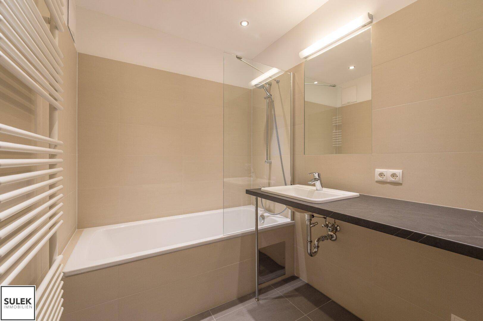 Modernes Badezimmer mit Badewanne, Einzelwaschbecken, beigen Wandfliesen und beheiztem Handtuchhalter.