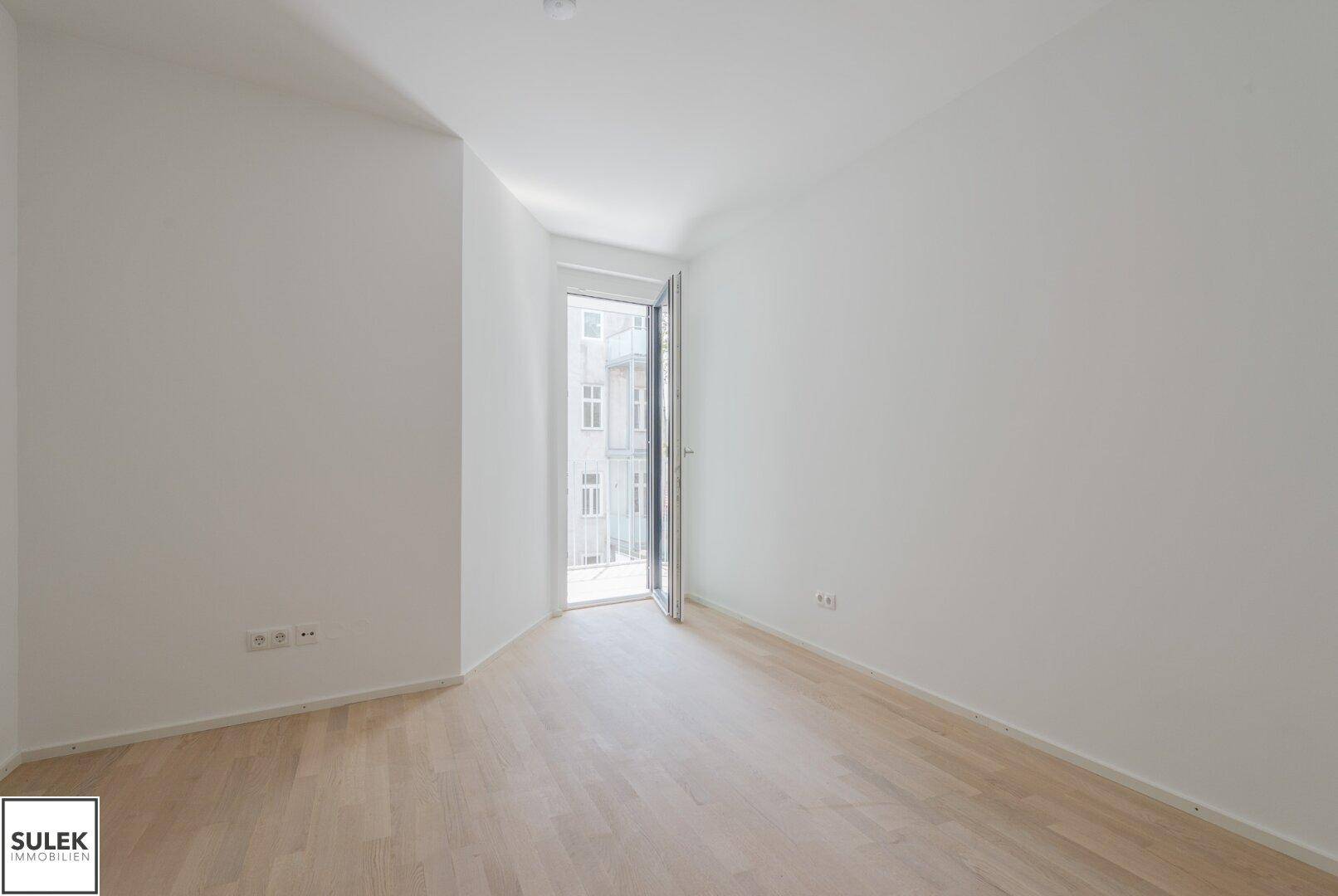 Leeres Zimmer mit hellem Holzboden, weißen Wänden und Zugang zu einem kleinen Balkon.