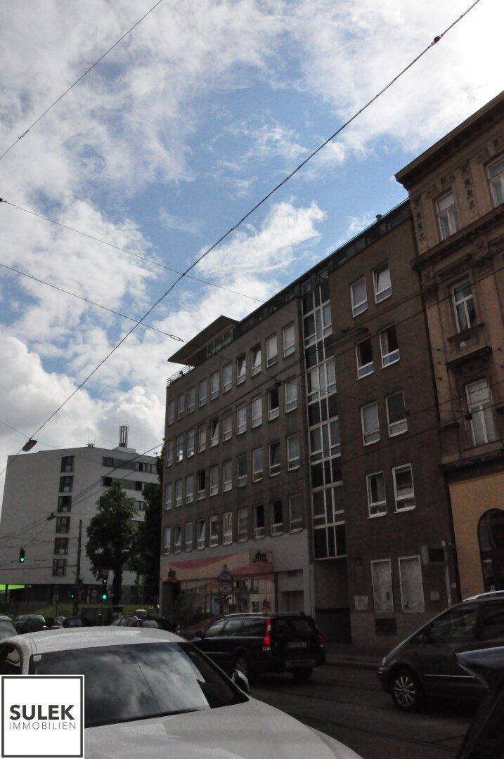 Eine belebte Stadtstraße mit verschiedenen Gebäuden und geparkten Fahrzeugen unter einem teilweise bewölkten Himmel.