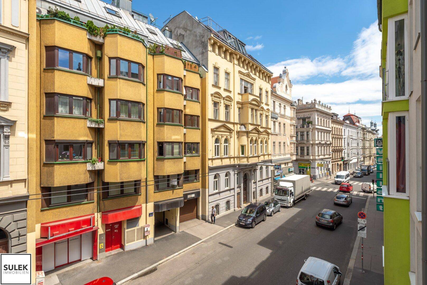 Panoramablick auf eine städtische Straße mit verschiedenen Gebäudefassaden und vorbeifahrenden Autos.
