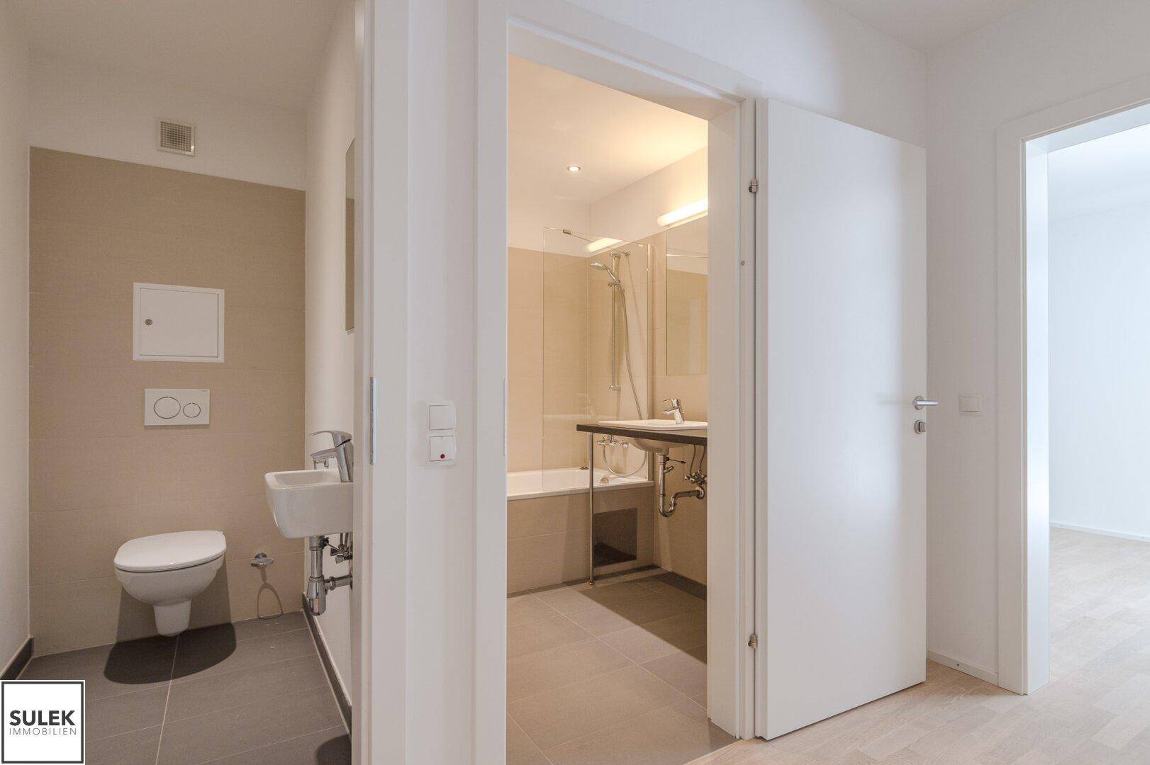 Modernes Badezimmer mit Wand-WC, kleinem Waschbecken und Zugang zu einem weiteren Badbereich, hell gefliest.