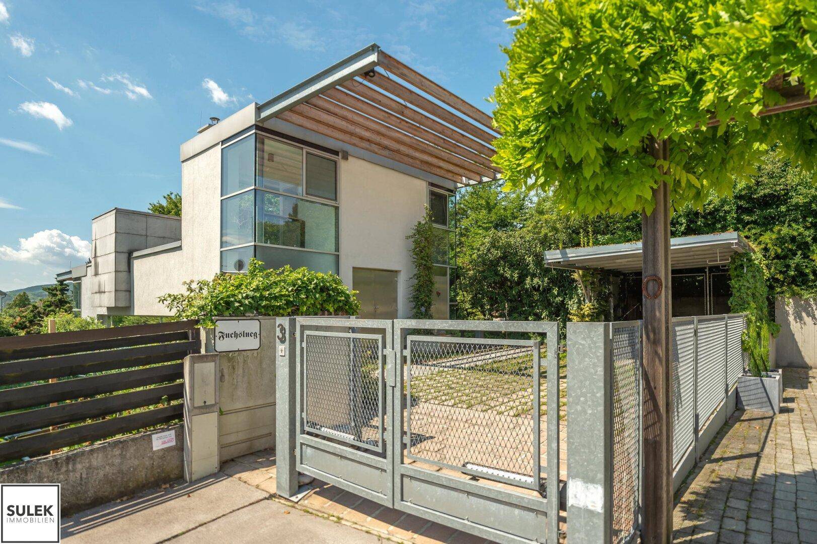 Moderne Architektur des Hauses mit Metallzaun und Einfahrt, umgeben von grüner Bepflanzung.