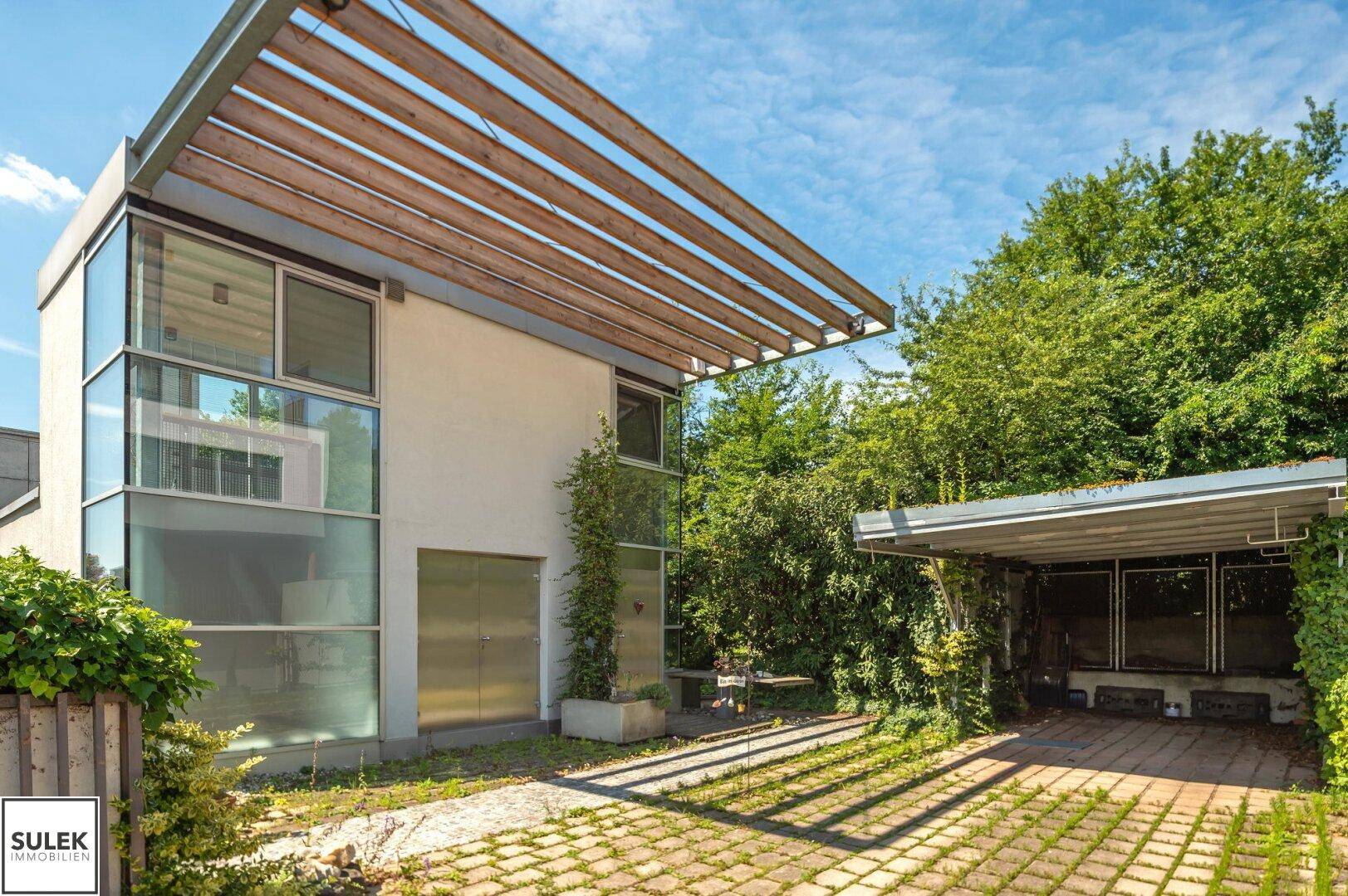 Seitenansicht des modernen Hauses mit Glasfassade und überdachtem Carport im Grünen.