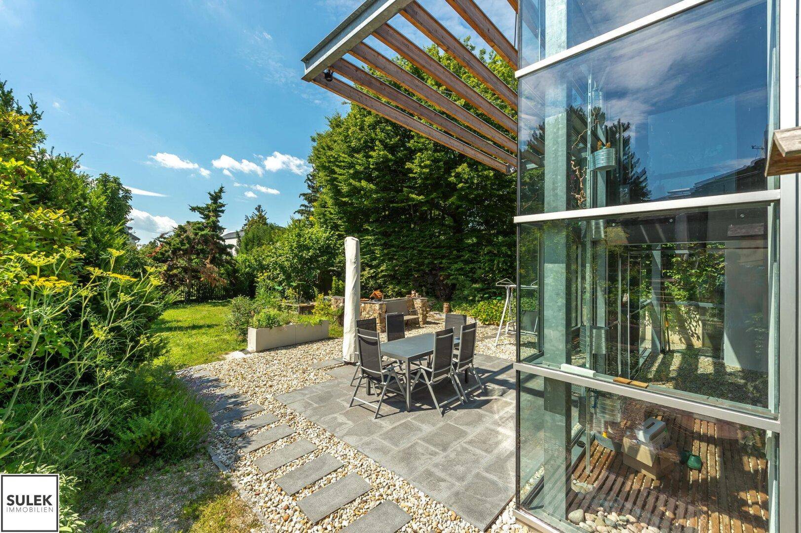 Sonniger Terrassenbereich mit Esstisch und Stühlen, umgeben von üppigem Grün und Kies.