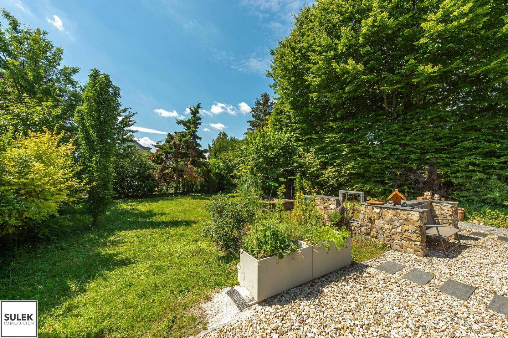 Großer, gepflegter Garten mit Rasenfläche, Hochbeet und Steinmauer unter blauem Himmel.