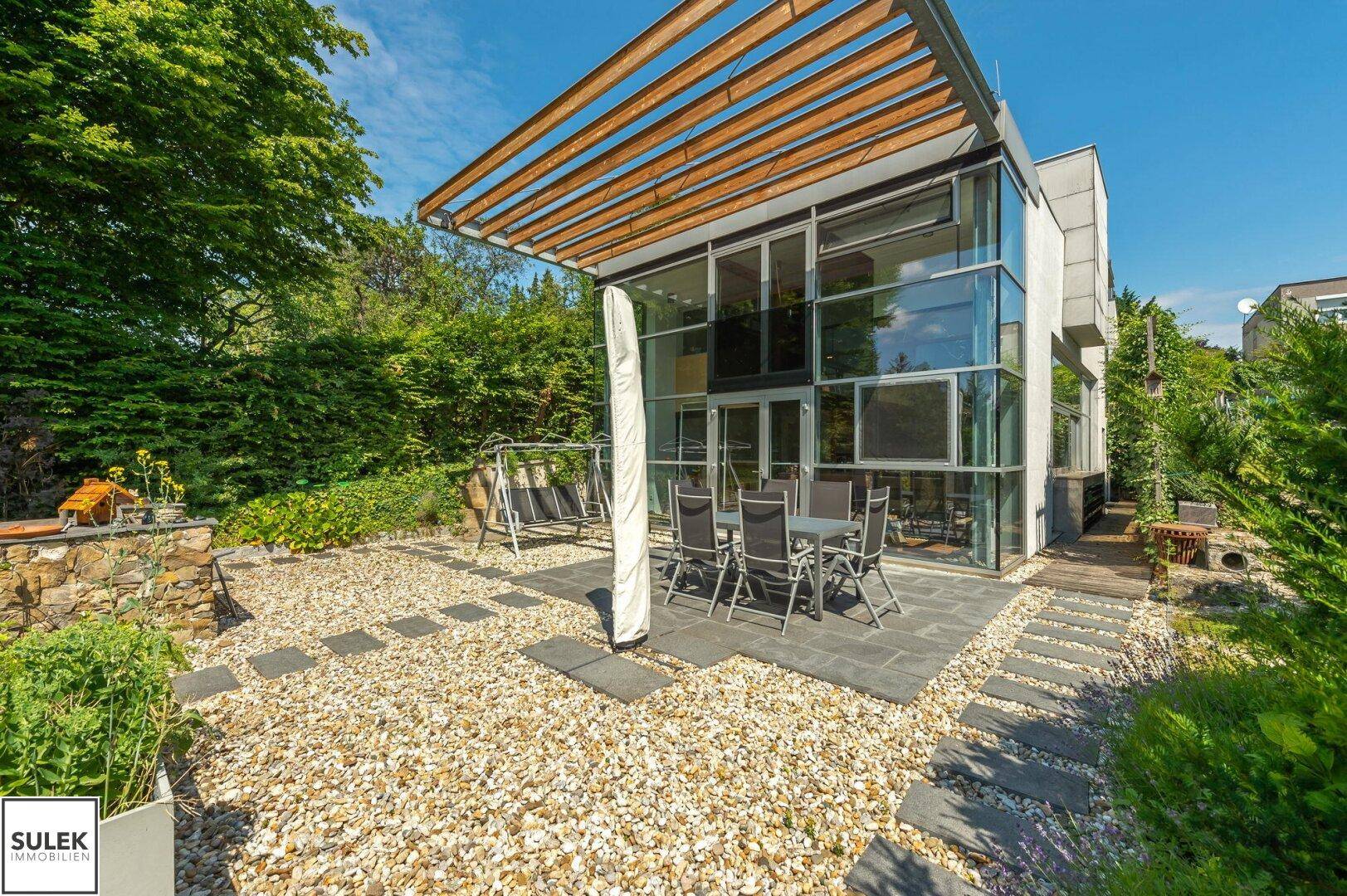 Sonniger Kiesgarten mit gepflasterter Terrasse, Esstisch und Schaukelstuhl vor moderner Glasfassade.