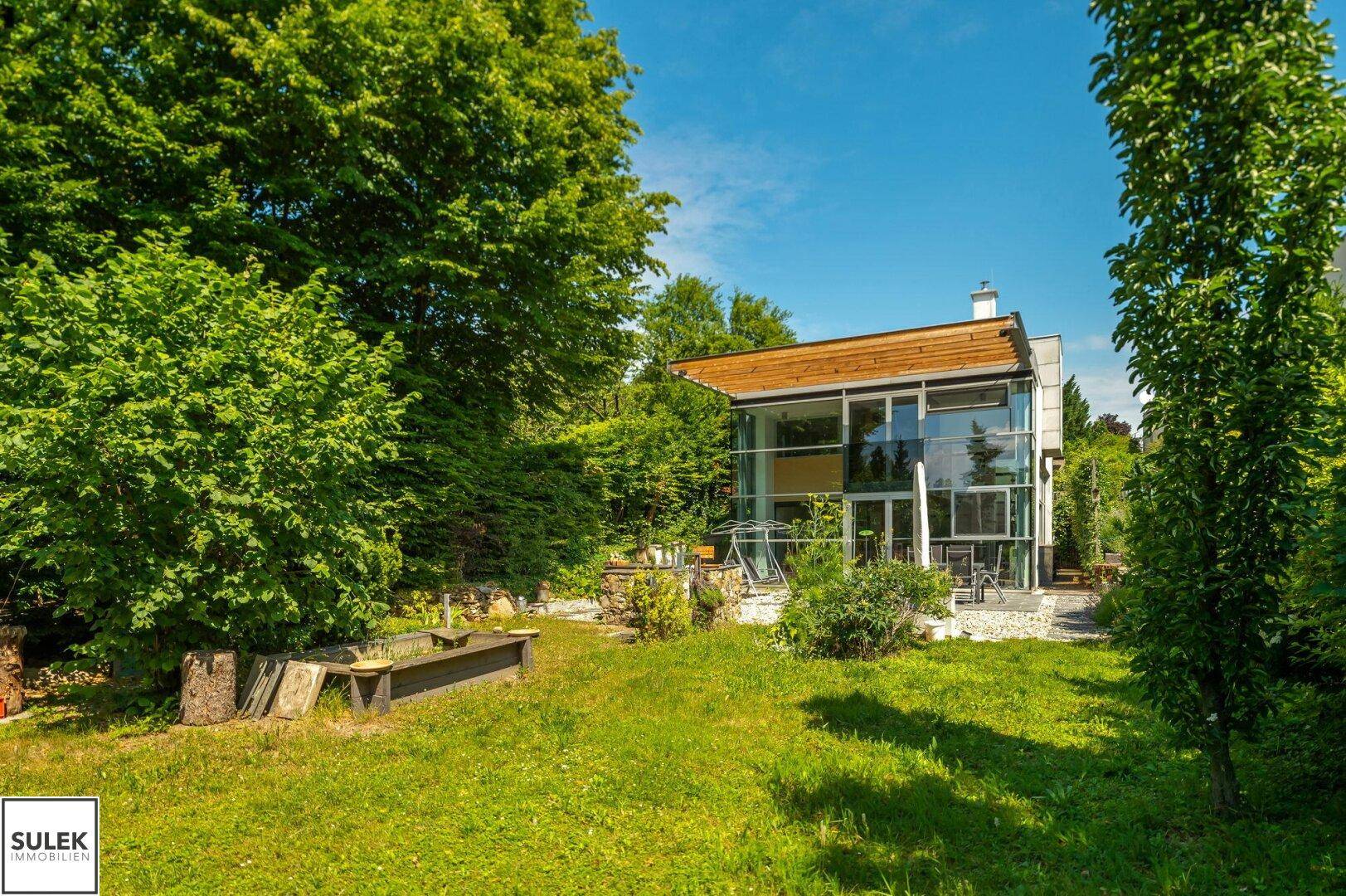 Weitläufiger Garten mit Rasen und Bäumen, im Hintergrund das moderne Haus mit Glasfassade.