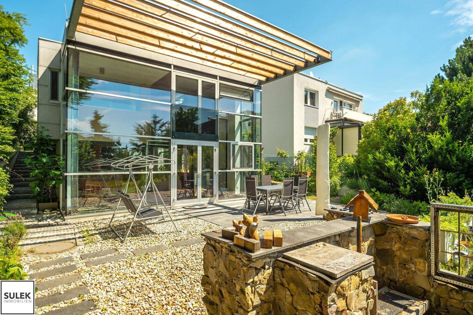 Sonniger Terrassenbereich mit Esstisch und dekorativer Steinmauer vor der Glasfassade des Hauses.