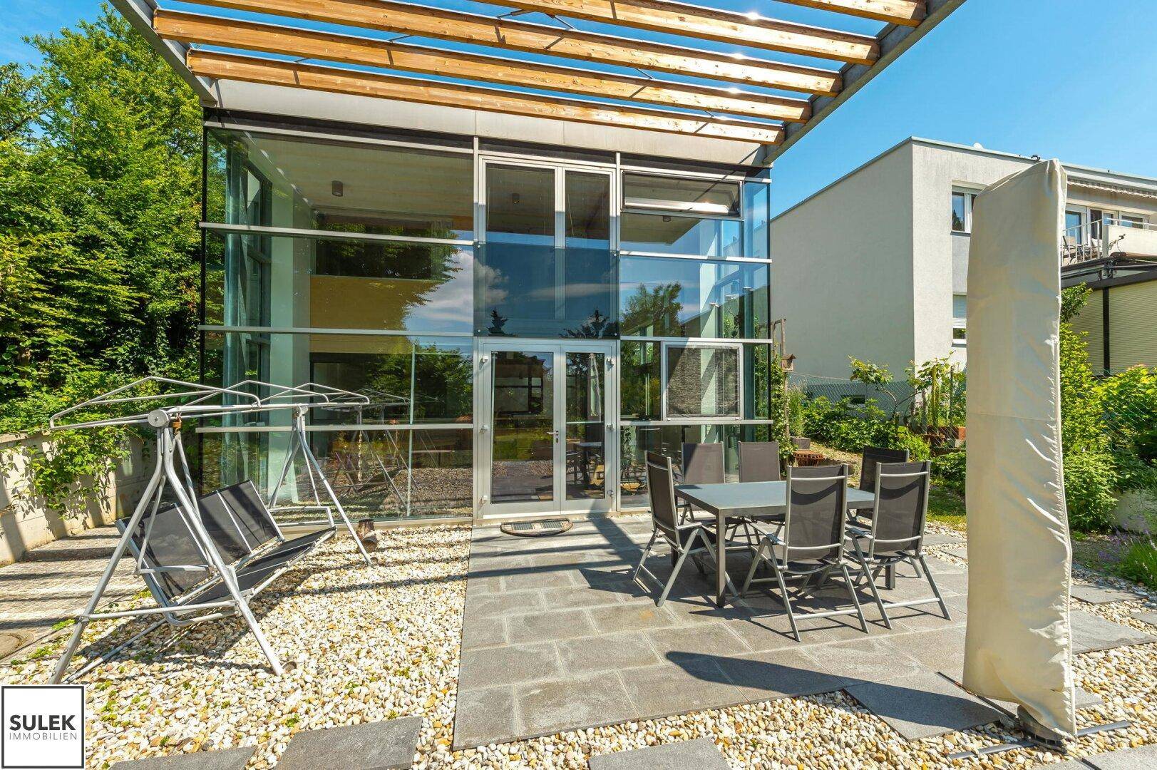 Möblierte Terrasse mit Esstisch und Sonnenschirm, umgeben von Kies und grüner Bepflanzung.