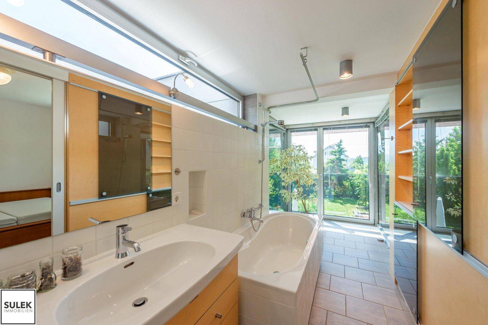 Modernes Badezimmer mit Badewanne, Doppelwaschbecken und großen Fenstern zum Garten.