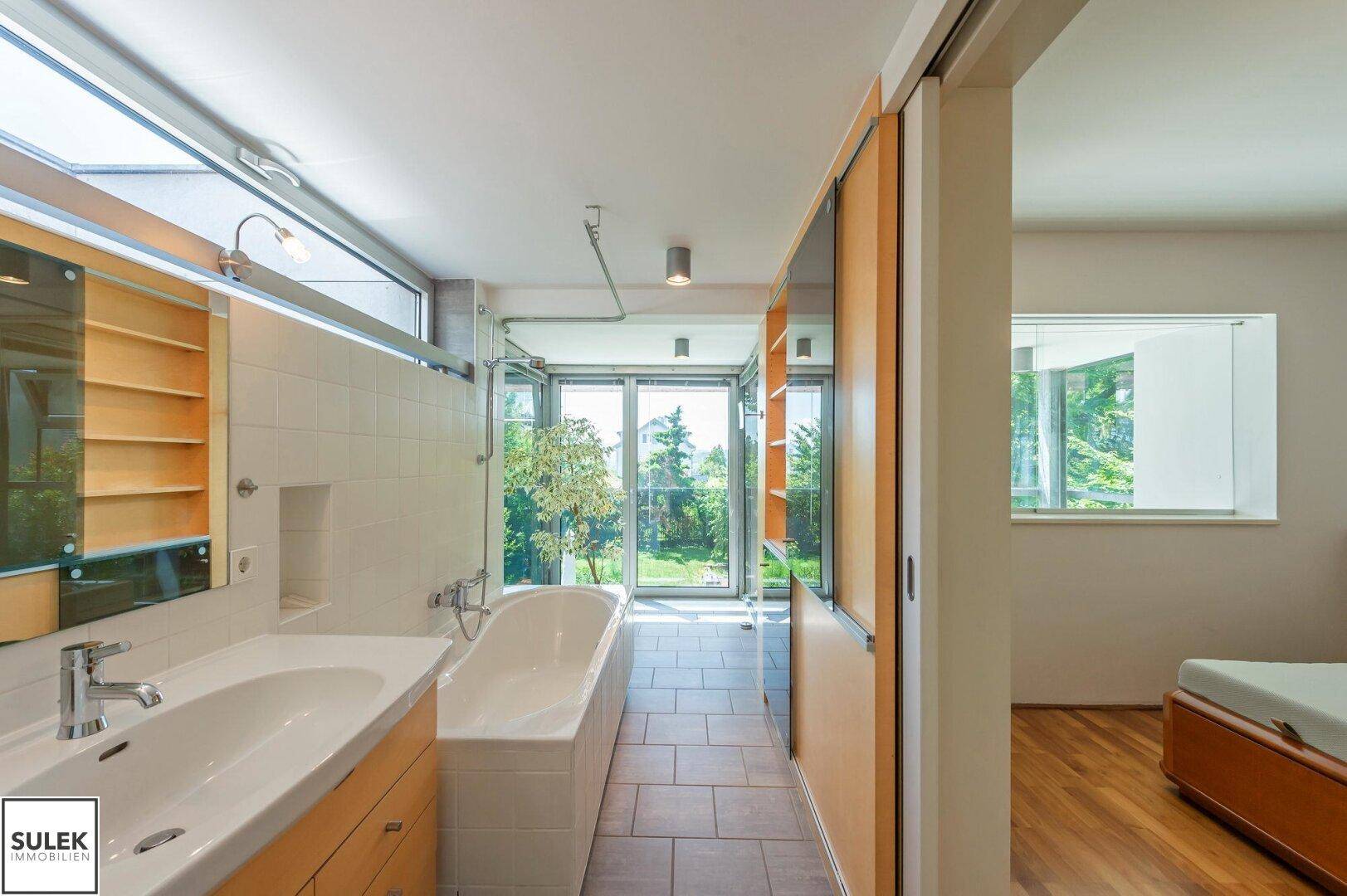 Helles Badezimmer mit Badewanne, Waschbecken und großen Fenstern mit Gartenblick, sehr einladend.
