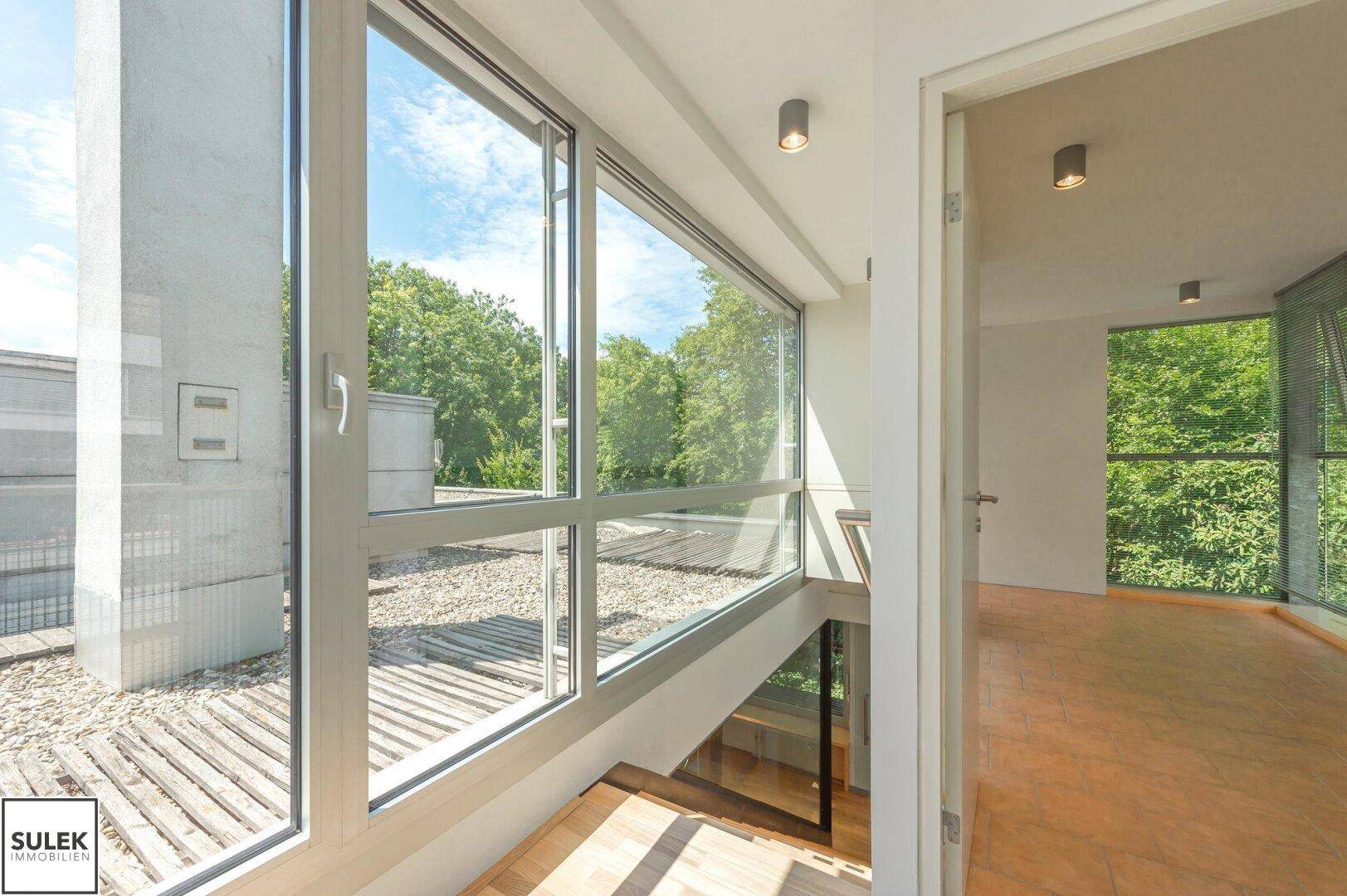 Heller Flur mit großen Fenstern, Holzboden und Blick auf die Dachterrasse und Bäume, sehr offen.