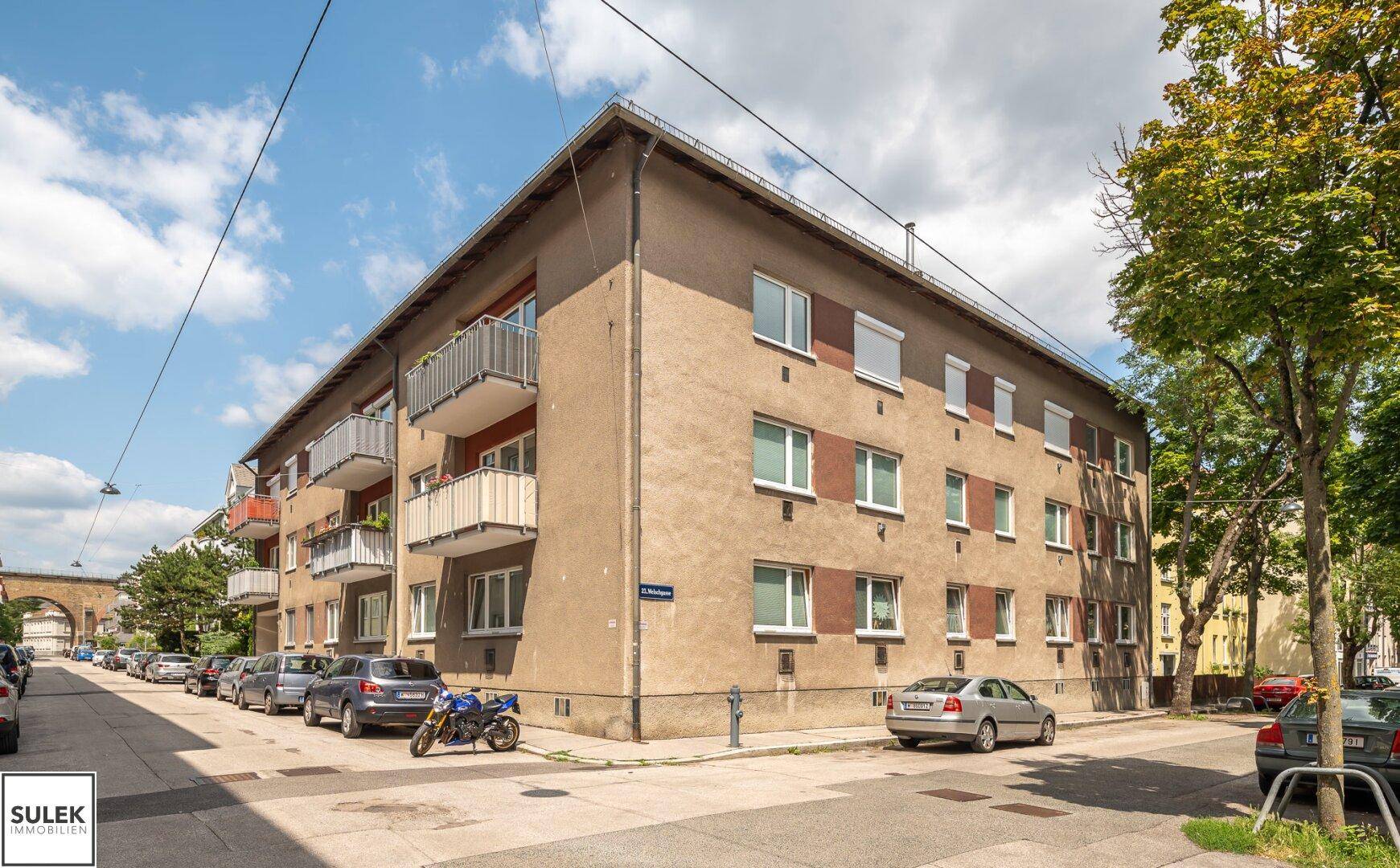 Mehrfamilienhaus mit Balkonen und geparkten Autos an einer sonnigen Straße.