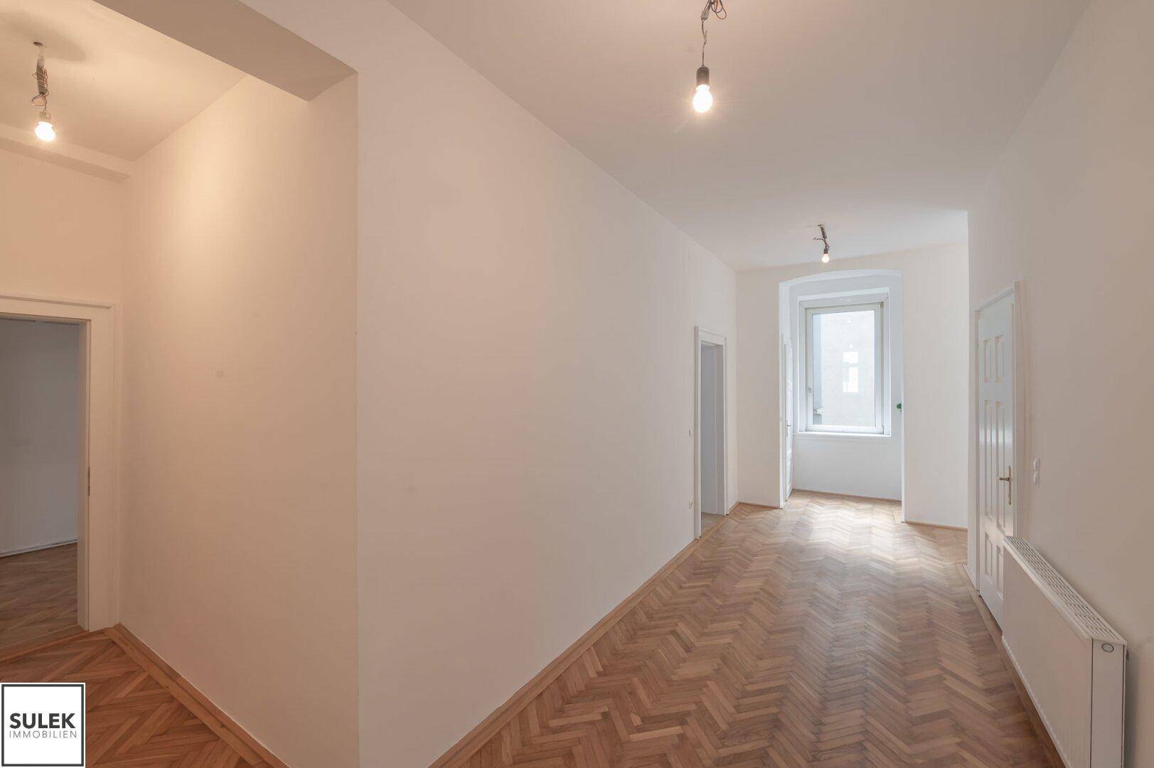Langer Flur mit Fischgrätparkett, weißen Wänden und einem Fenster am Ende.