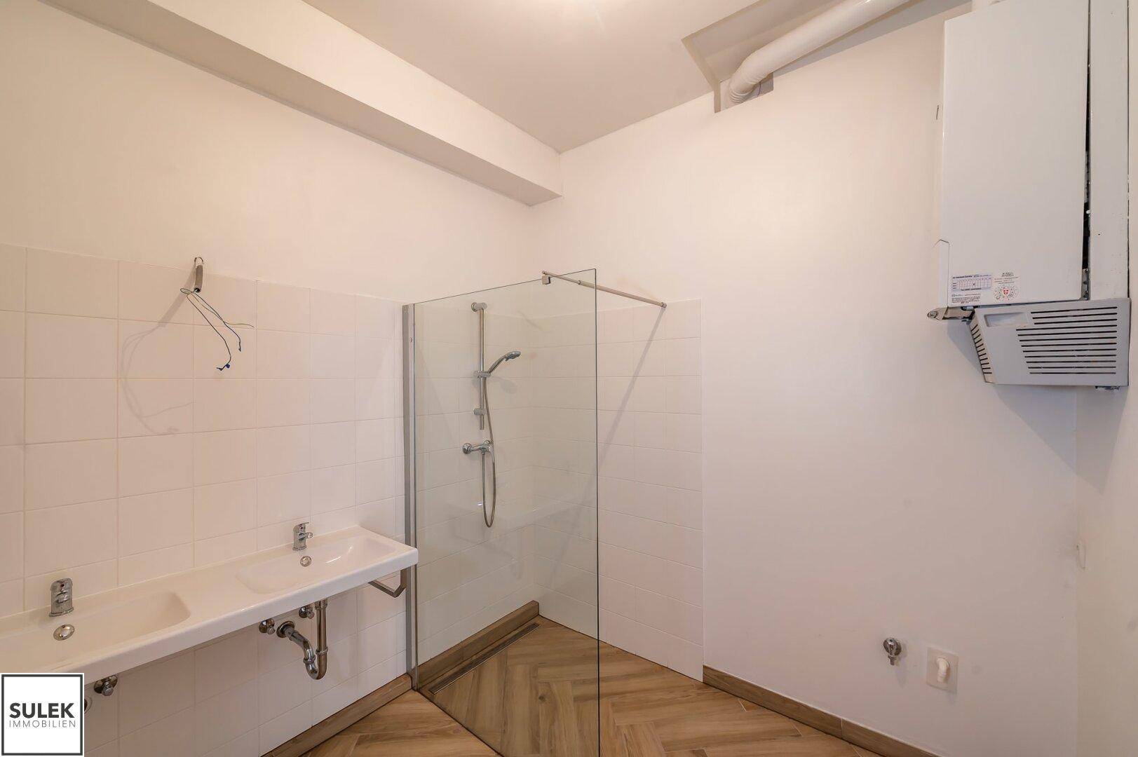 Badezimmer mit begehbarer Dusche, zwei Waschbecken und einem Warmwasserbereiter an der Wand.