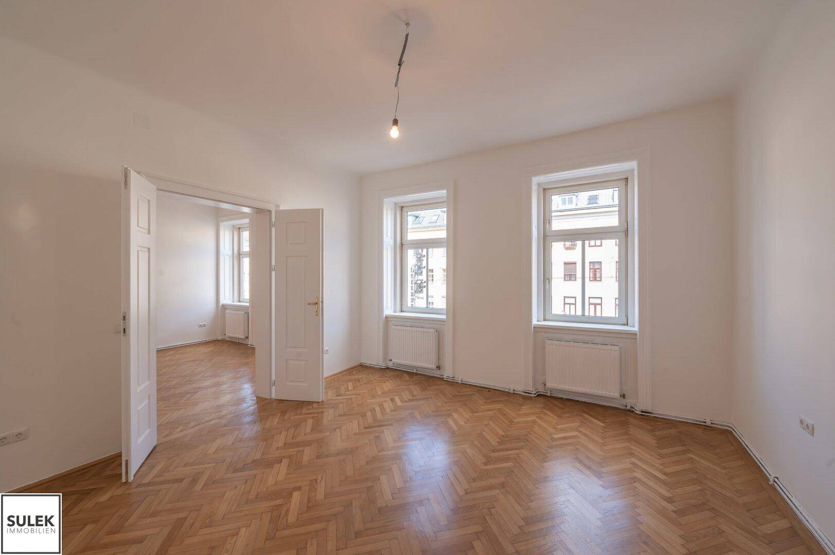 Geräumiges Zimmer mit Fischgrätparkett, hohen Fenstern und Blick auf die Stadt.