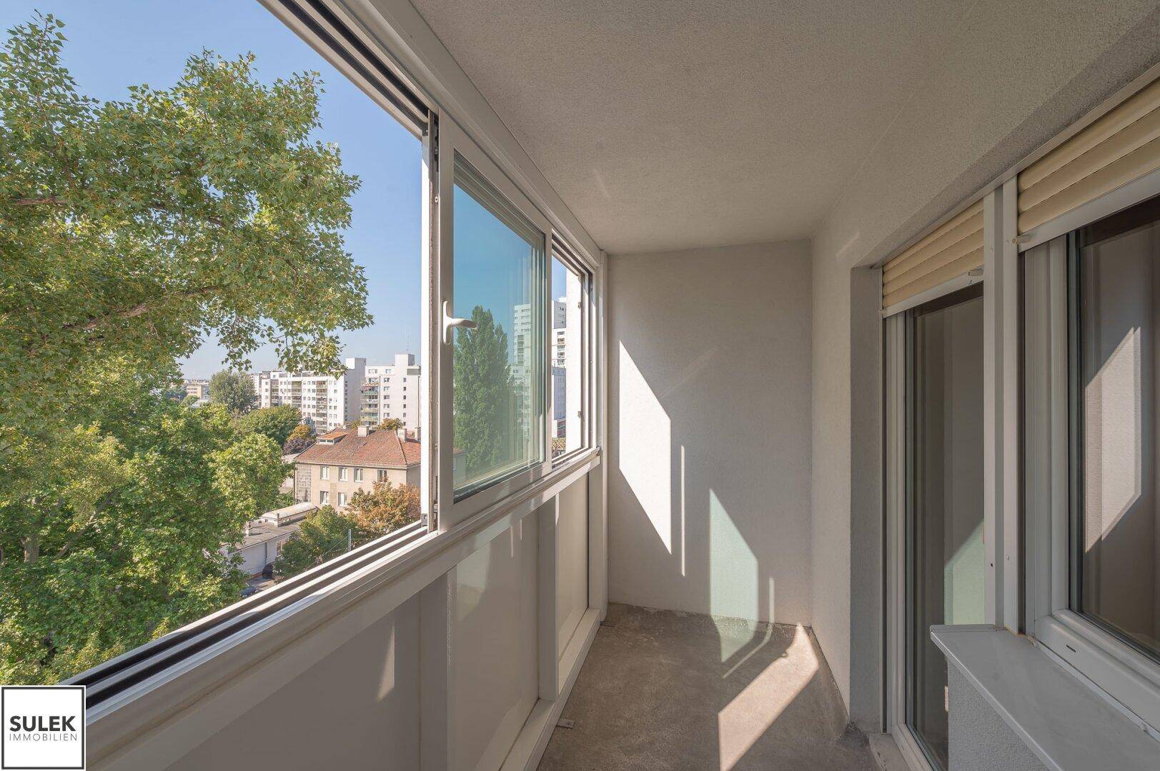 Verglaste Loggia mit weitem Blick über die Stadt und grüne Bäume.