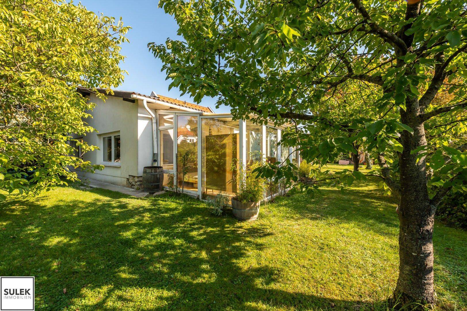 Idyllischer Garten mit altem Baumbestand und einem hellen Wintergarten am Haus bei Sonnenschein.