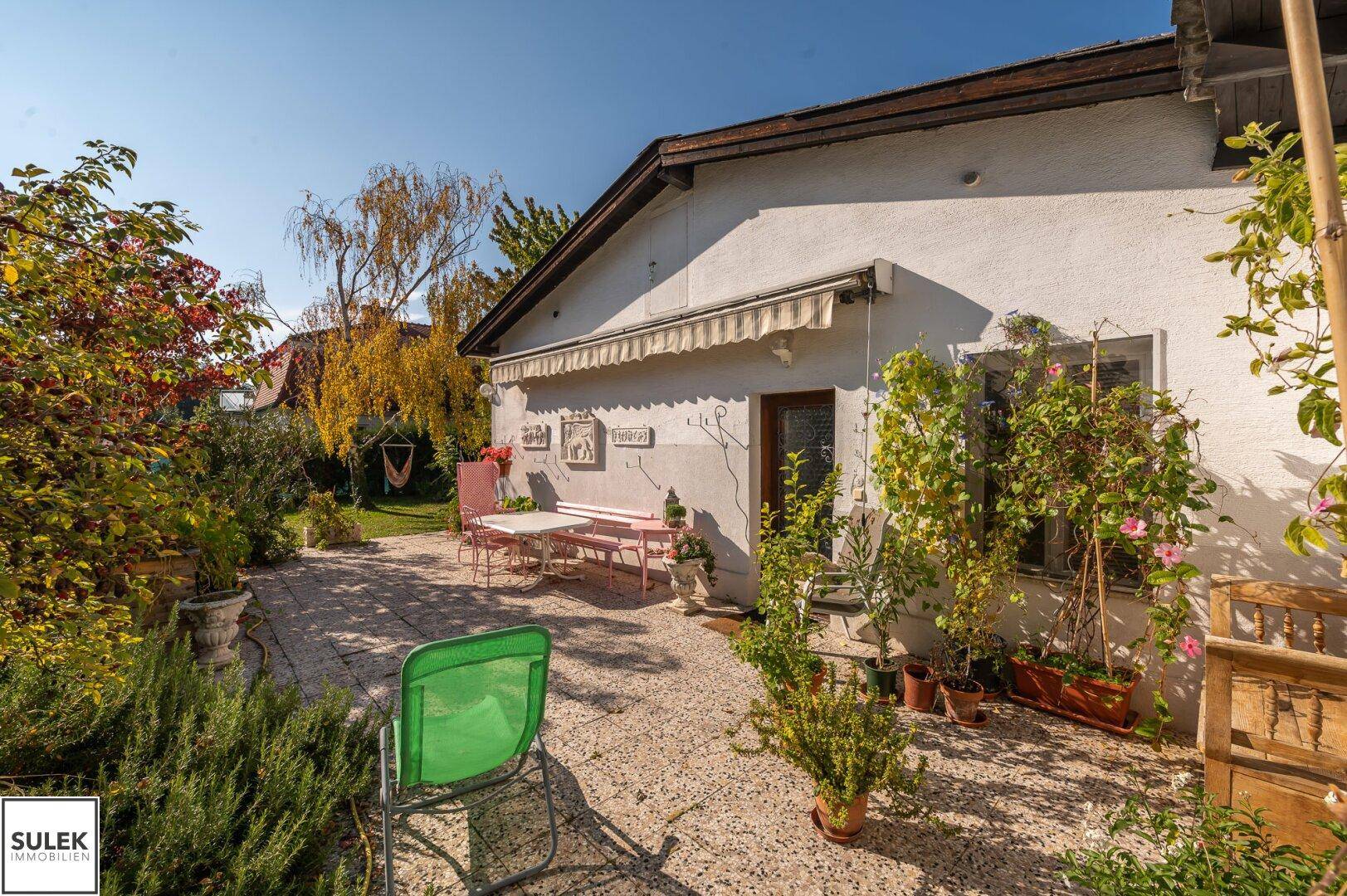 Sonniger Terrassenbereich mit Gartenmöbeln und Blumen, umgeben von üppigem Grün.