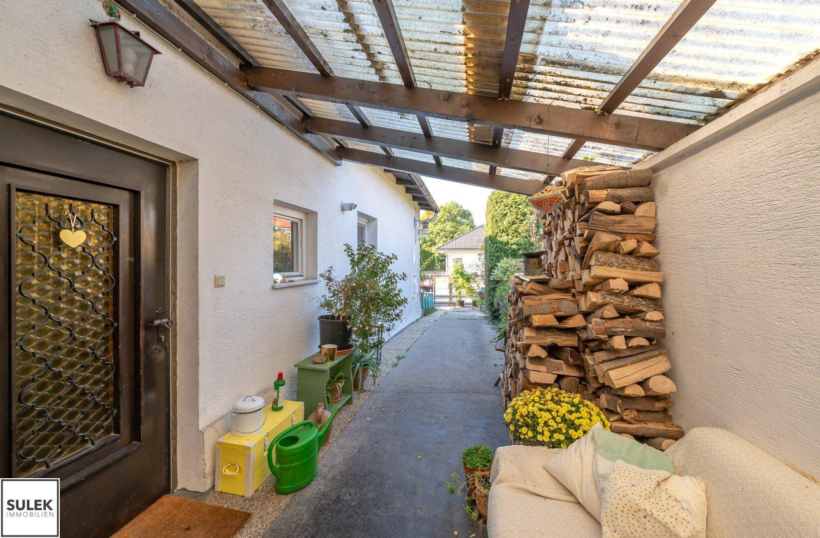 Überdachter Außenbereich mit Holzlager, Sitzbank und Zugang zum Haus.