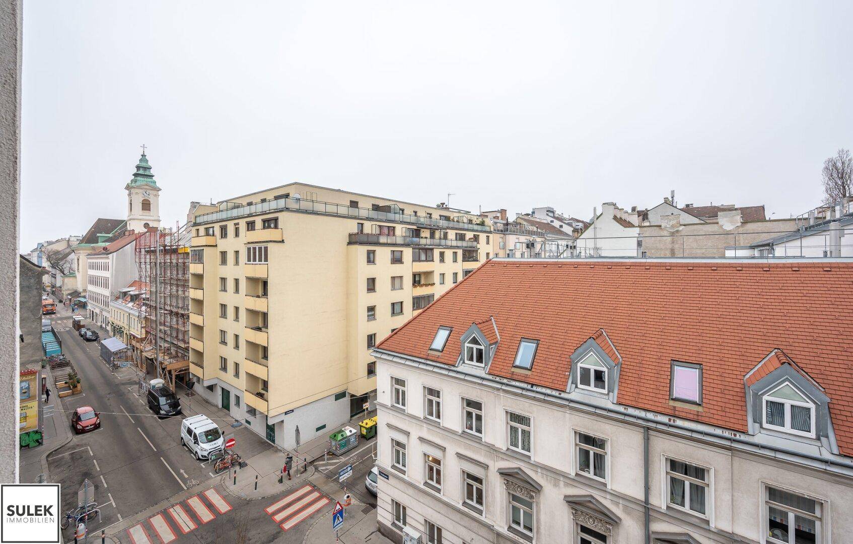 Blick aus dem Fenster auf eine belebte Straße mit umliegenden Gebäuden und Dächern der Stadt.