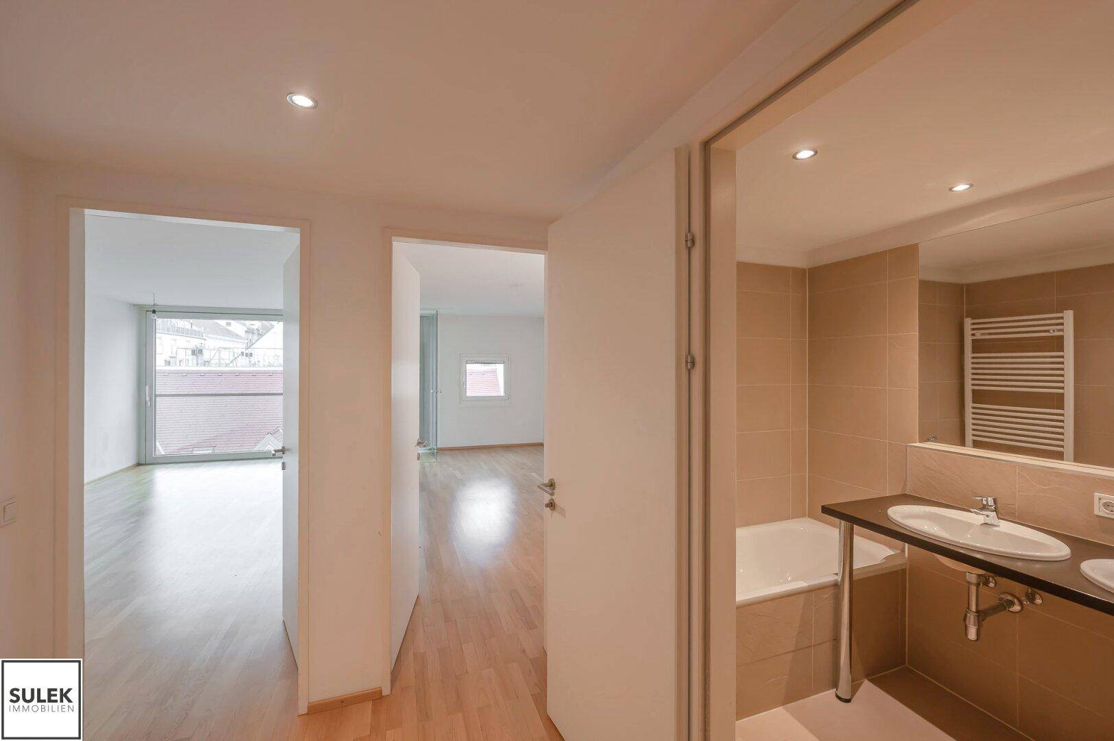 Flur mit Holzboden, der zu zwei Zimmern führt, darunter ein Badezimmer mit Badewanne und Doppelwaschbecken.