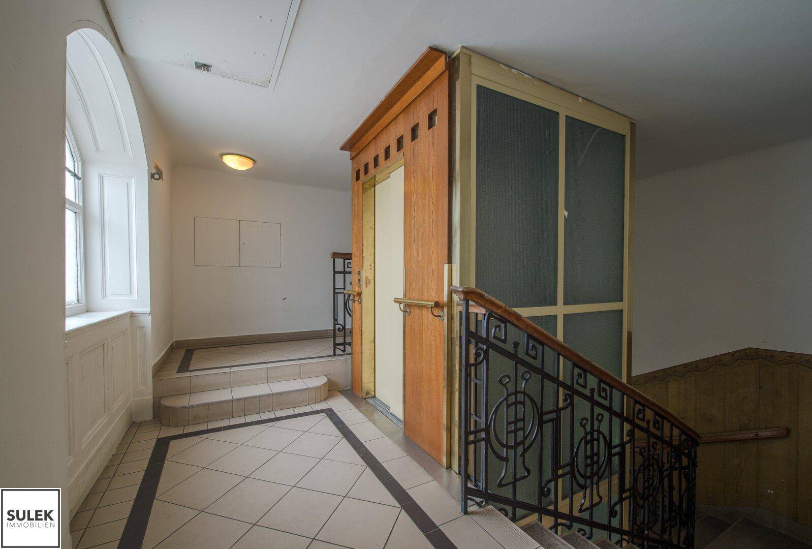 Historisches Treppenhaus mit Aufzug, gefliestem Boden und schmiedeeisernem Geländer.