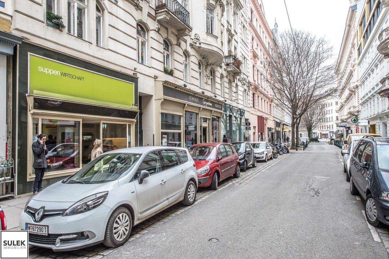 Stadtstraße mit parkenden Fahrzeugen und Geschäften im Erdgeschoss historischer Gebäude.