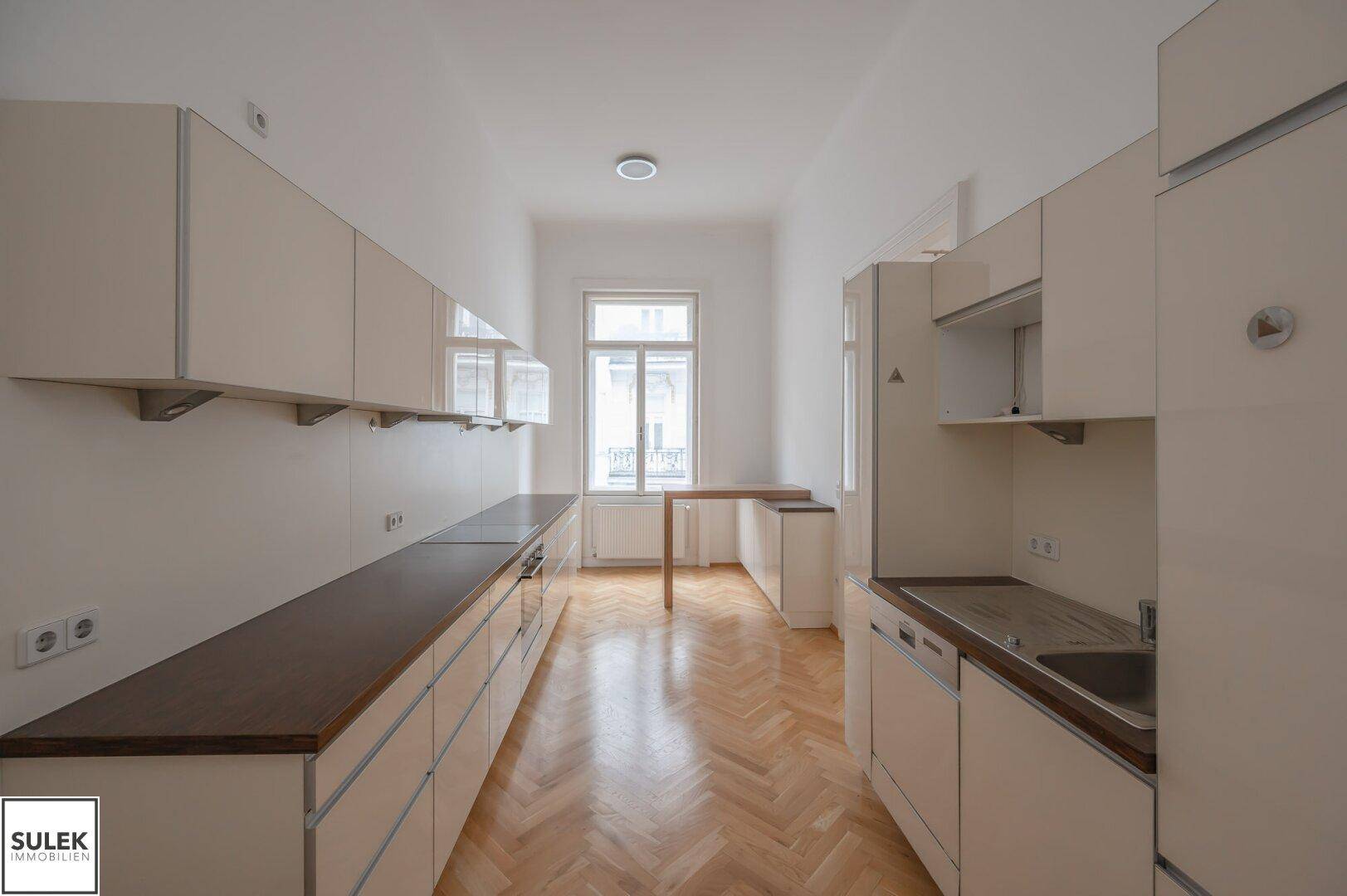 Lange Küche mit Einbauschränken, dunklen Arbeitsplatten und Fenster am Ende des Raumes.