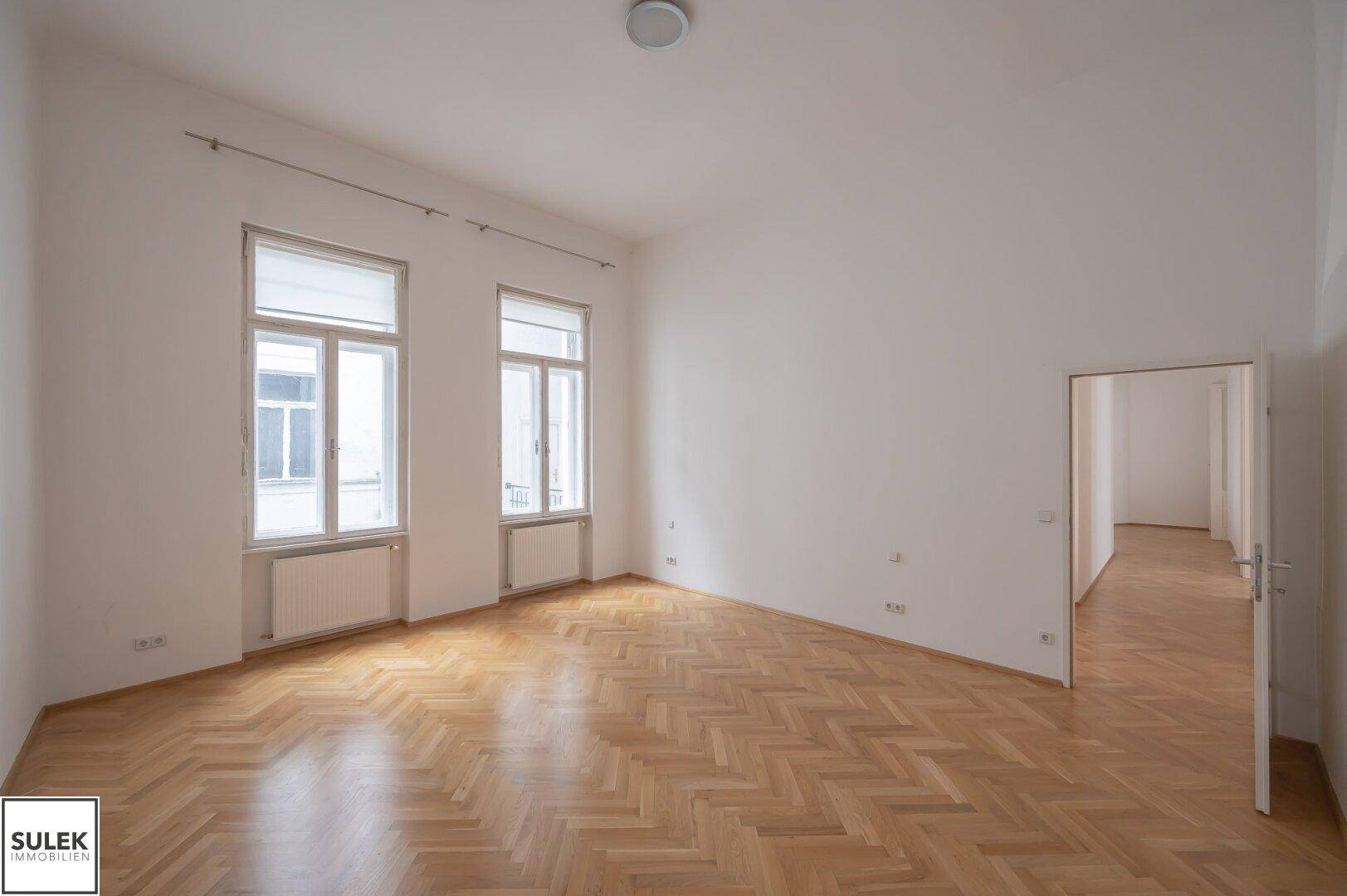 Heller Raum mit Fischgrätparkett, zwei Fenstern und Zugang zu einem weiteren Zimmer.