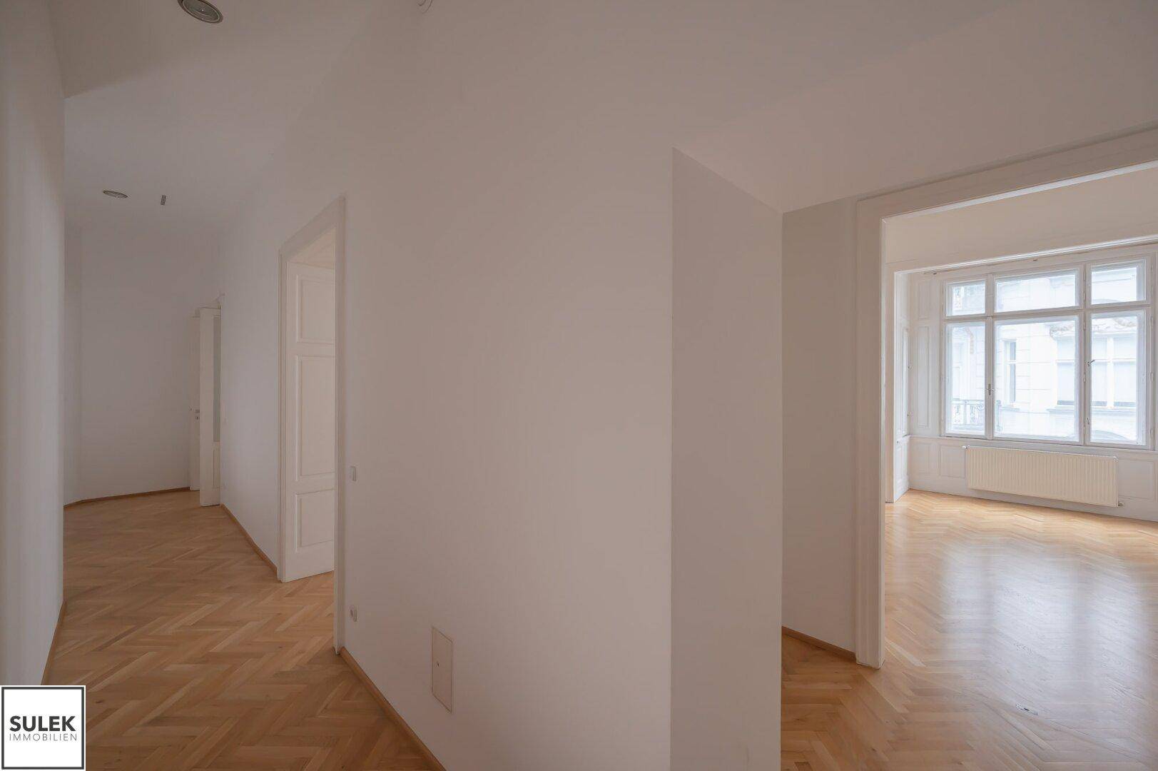 Gangbereich mit Fischgrätparkett, der zu einem hellen Zimmer mit Fenstern führt.