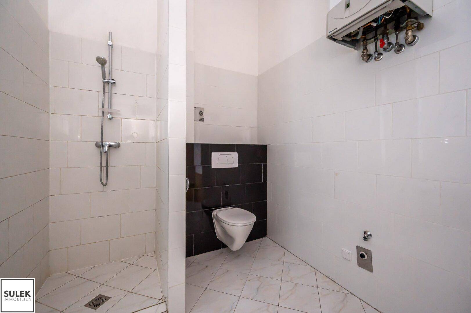 Badezimmer mit Duschbereich, Wand-WC und Warmwasserbereiter an der Wand.